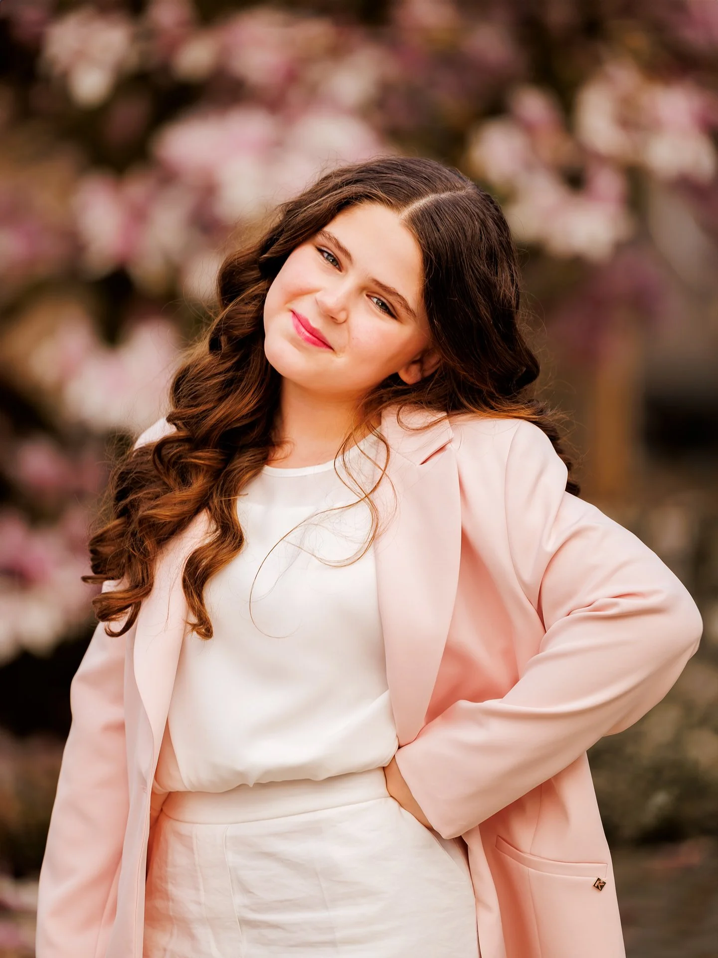 Portrait of a young woman with wavy brown hair, smiling softly, wearing a light pink blazer over a white top, standing outdoors with blurred pink and white flowers in the background.