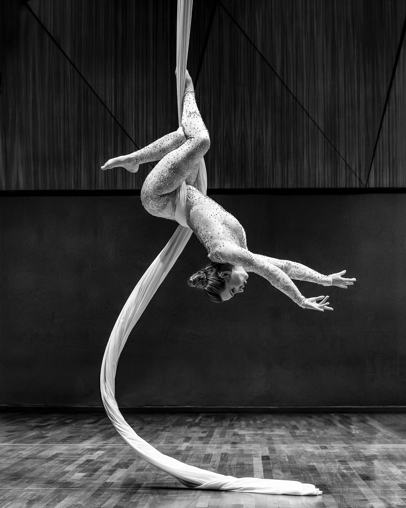 A female aerial silk performer dressed in a sparkly, fitted costume poses upside down on white aerial silks hanging from the ceiling against a dark background, true aerial art in motion.