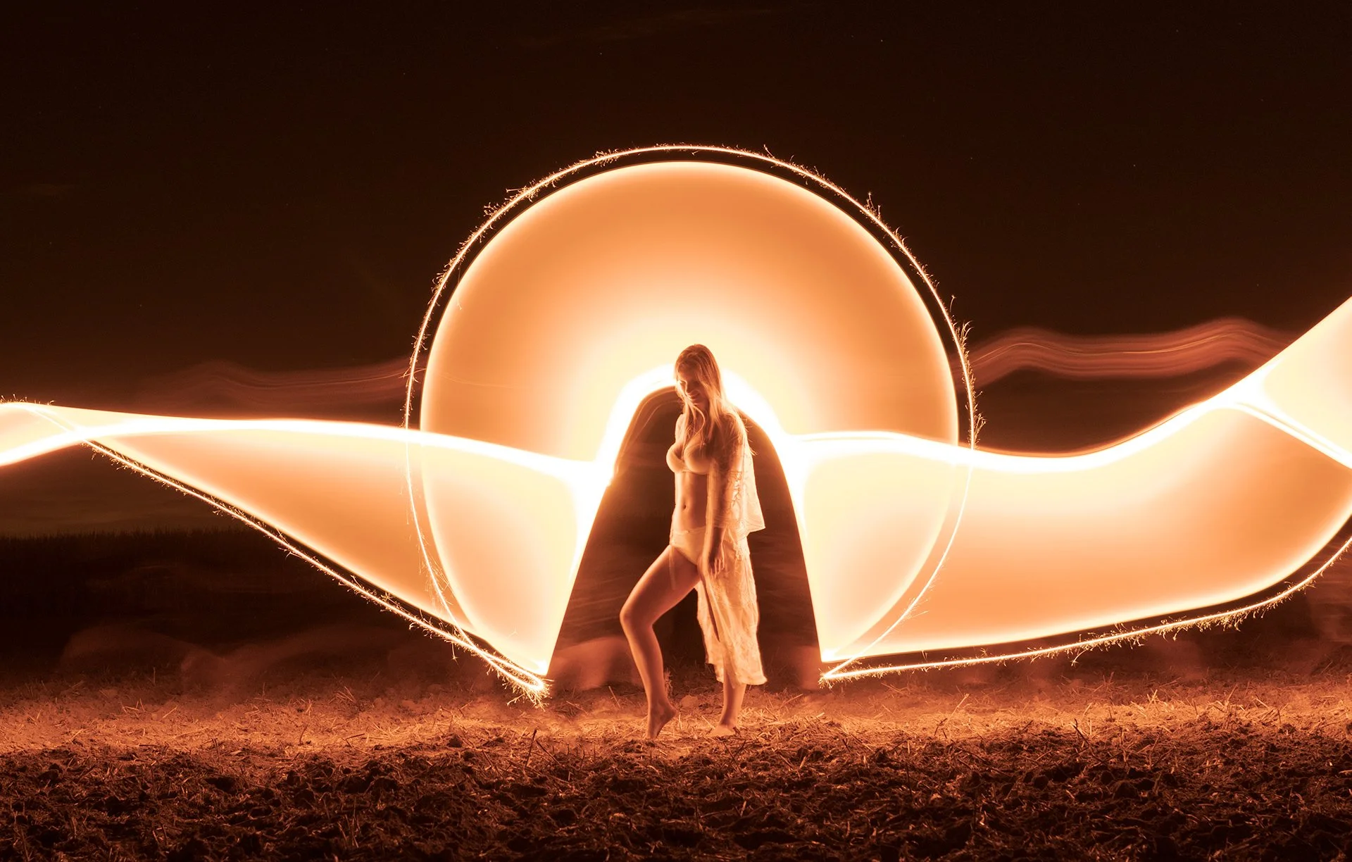 A light art photo of a woman standing in front of a large, illuminated, circular light art at night, with light trails creating a heart shape.