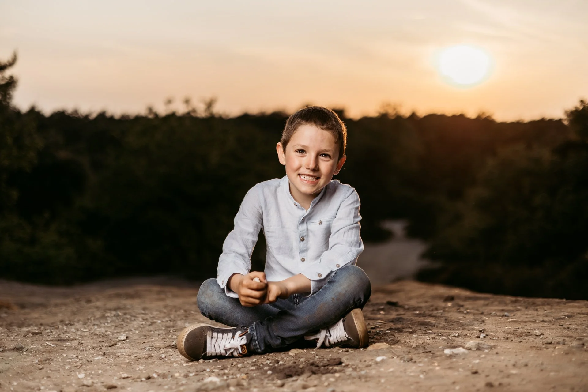 A young boy sitting cross-legged outdoors on a dirt path during sunset, smiling at the camera.