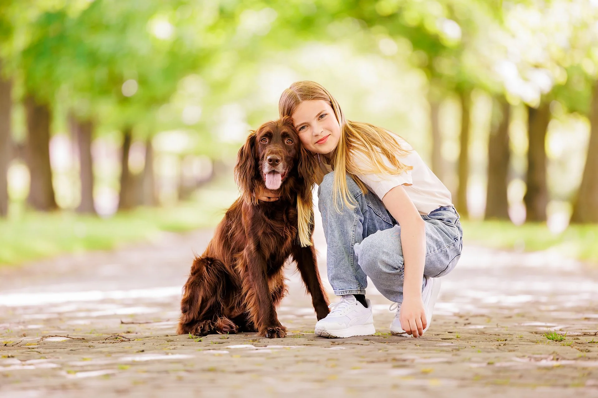 A young girl crouching next to a large brown dog on a paved pathway in a park with green trees in the background.