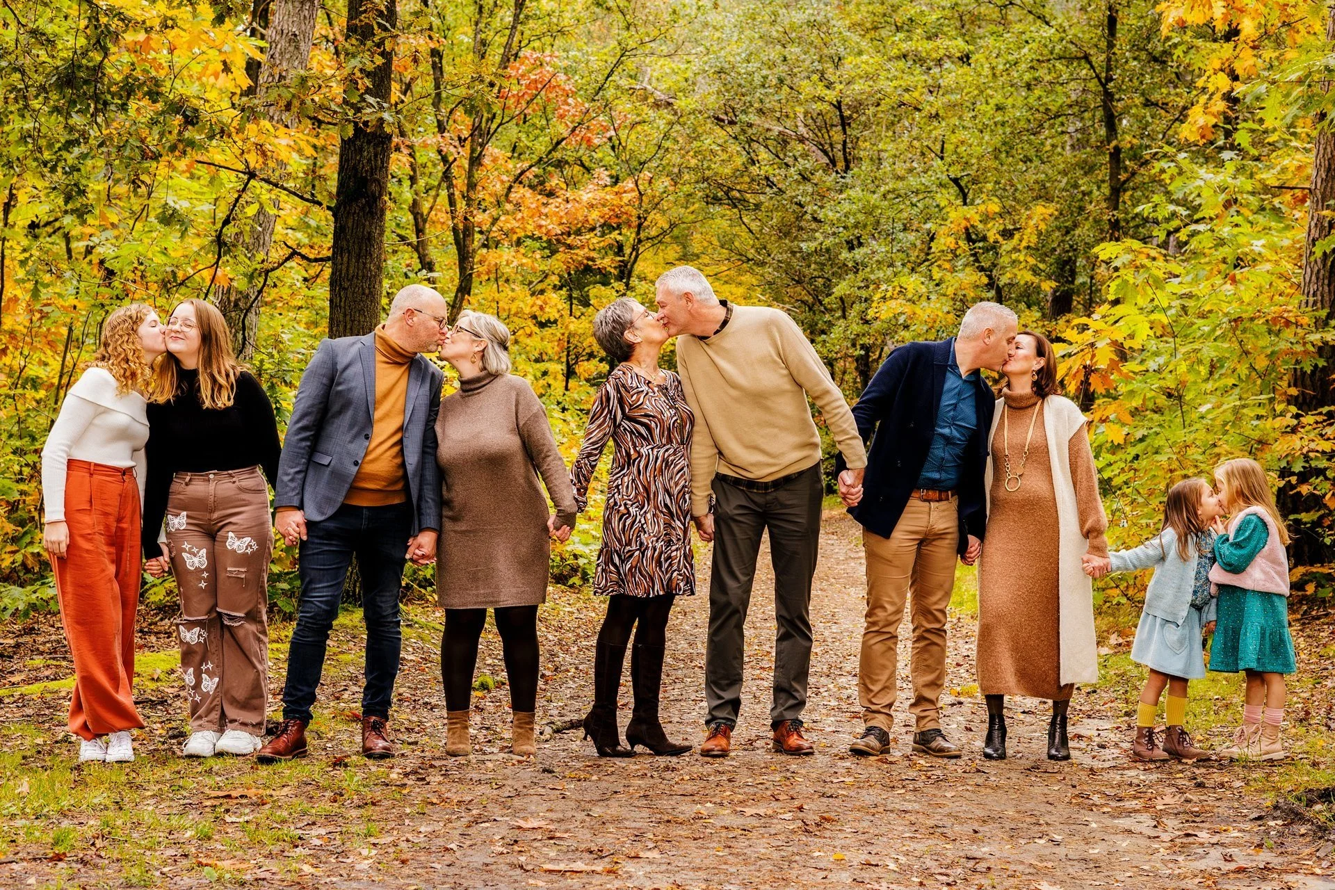 Multiple generations of a family, including children, parents, grandparents, and great-grandparents, holding hands and kissing in a forest during autumn.