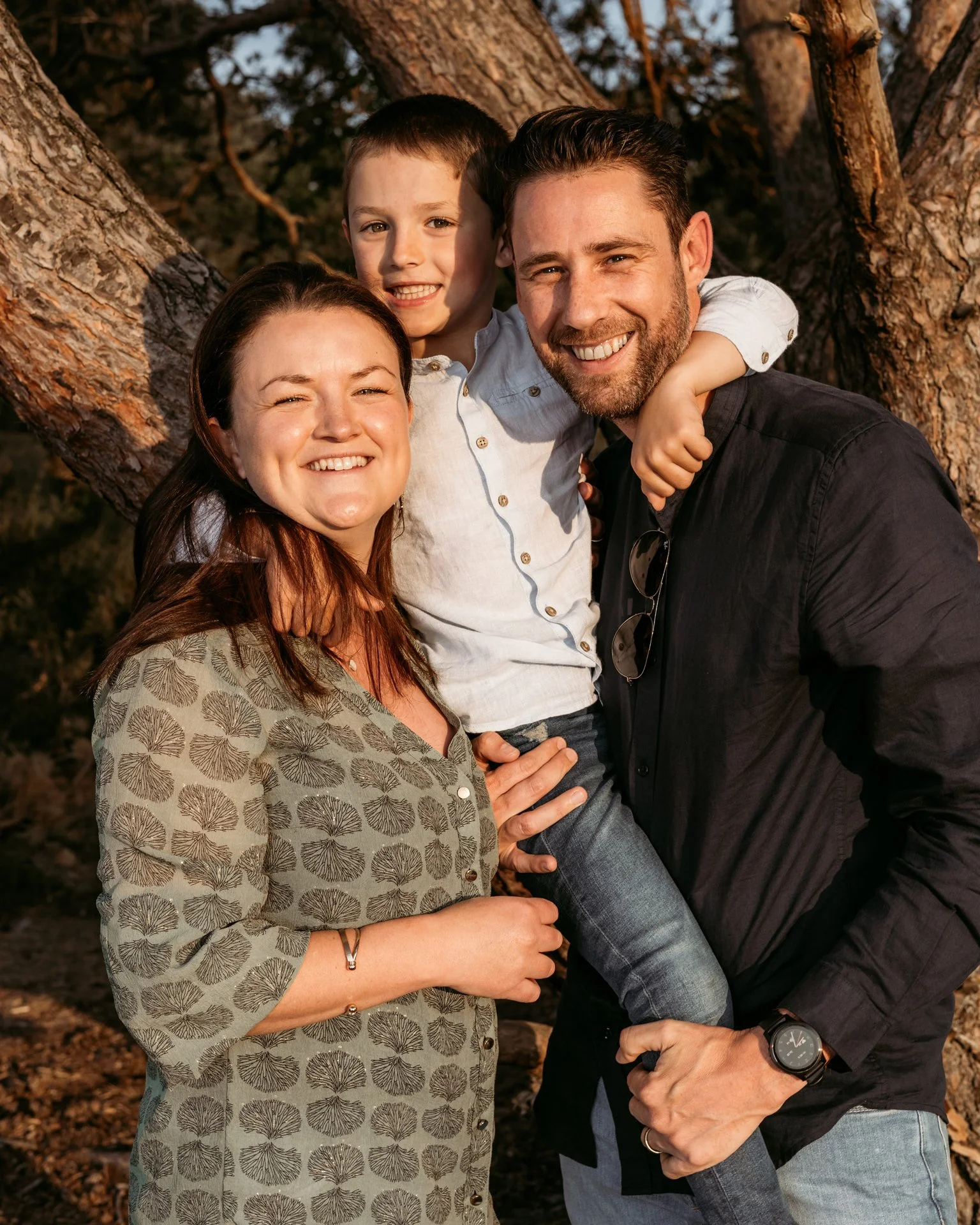 Happy family of three posing outdoors near a large tree during sunset, smiling at the camera.