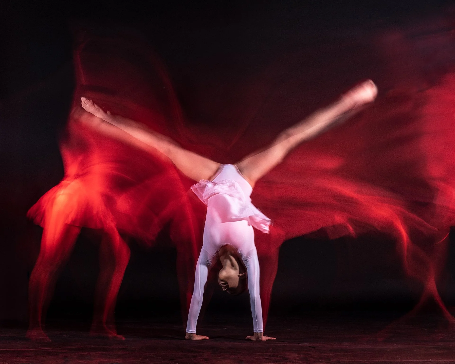 A dancer in a white costume performs a handstand with her arms on the floor and her legs extended outward. Red light painting creates motion blur effects around her