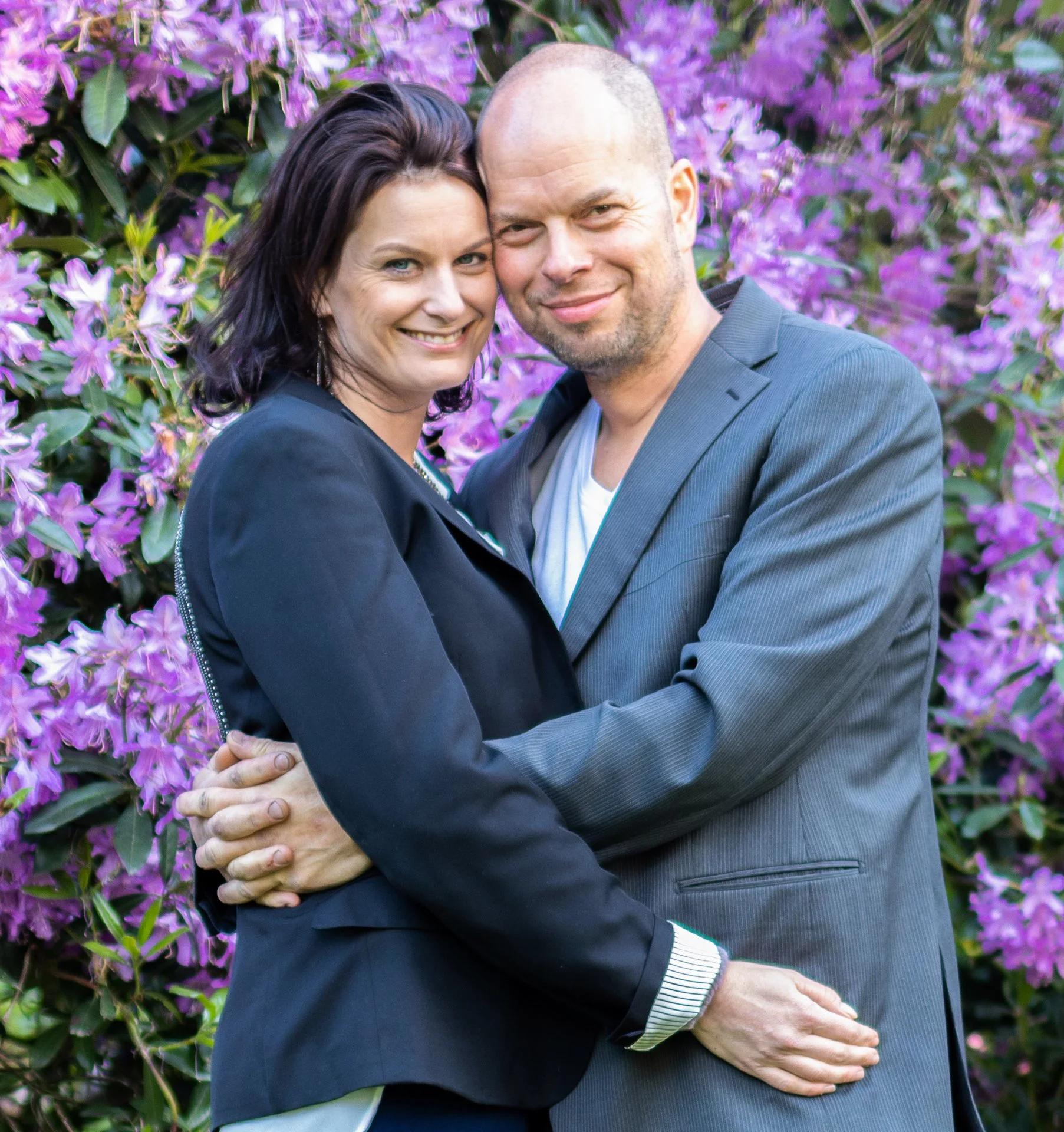 A smiling couple standing close together, embracing each other outdoors in front of purple flowers.