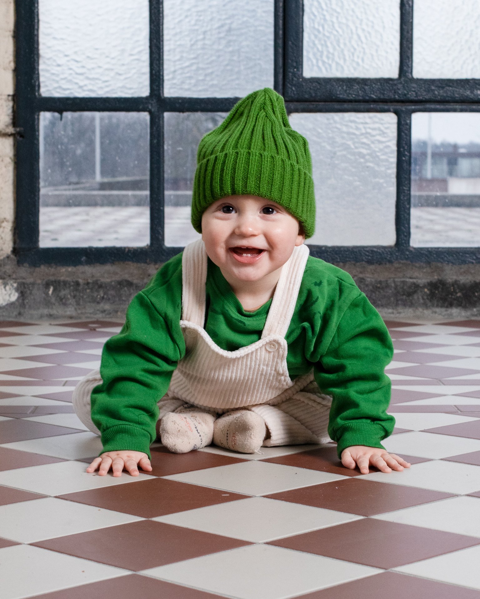 A smiling young child with a green knit hat, green shirt, beige overalls, and socks, crawling on a checkered tile floor in front of a window.