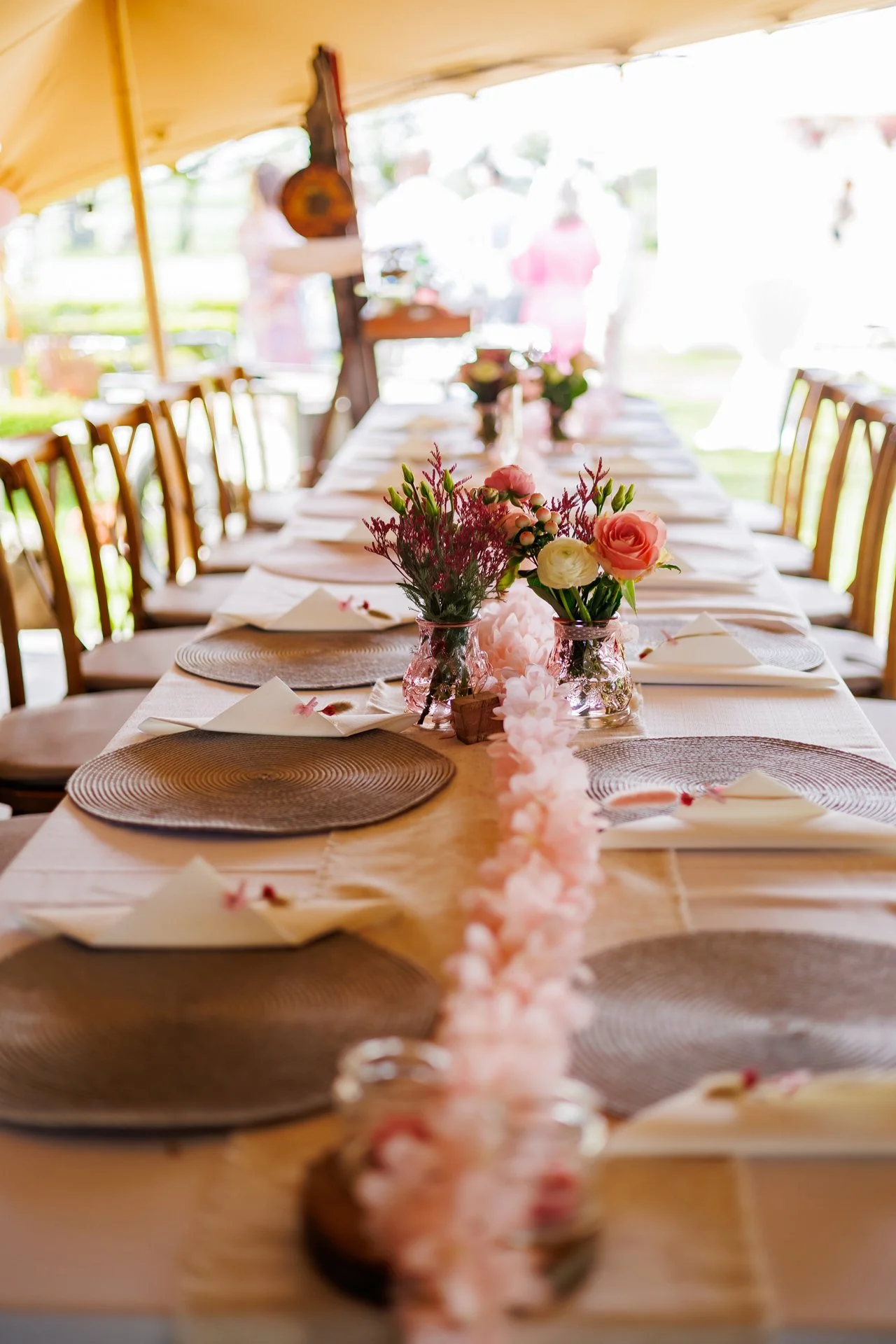 Long dining table with floral centerpieces and place settings under a tent at an outdoor event.