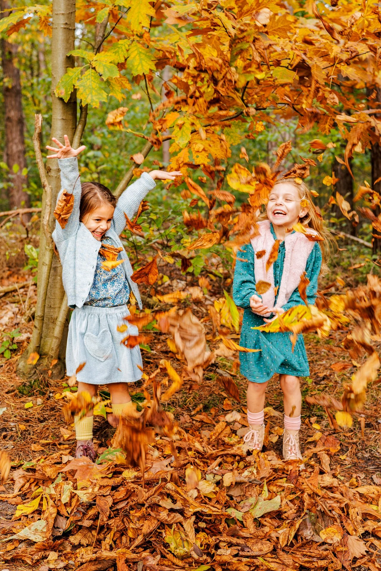 Two young girls playing and throwing autumn leaves in a forest with trees and colorful fall foliage.