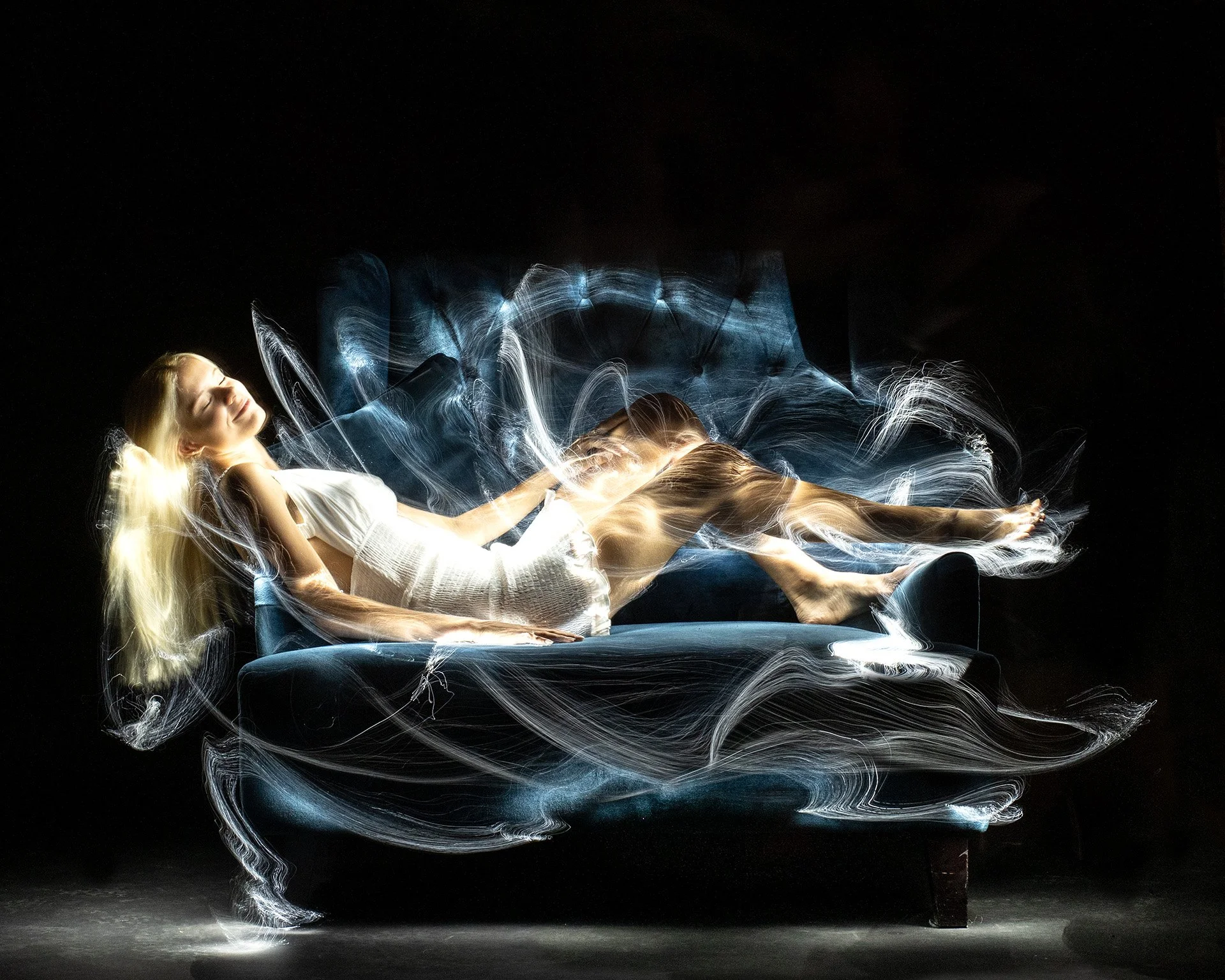Light painting around a reclining woman with long blonde hair in a white dress on a black couch against a dark background, creating an ethereal, motion-filled scene.