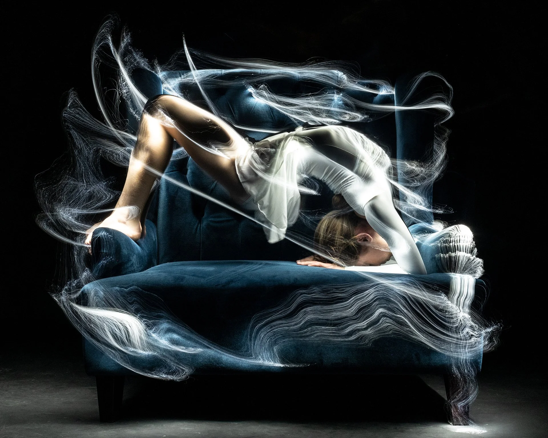 A person performing a backbend on a chair, with swirling light effects surrounding them in a dark setting. Light art photography.