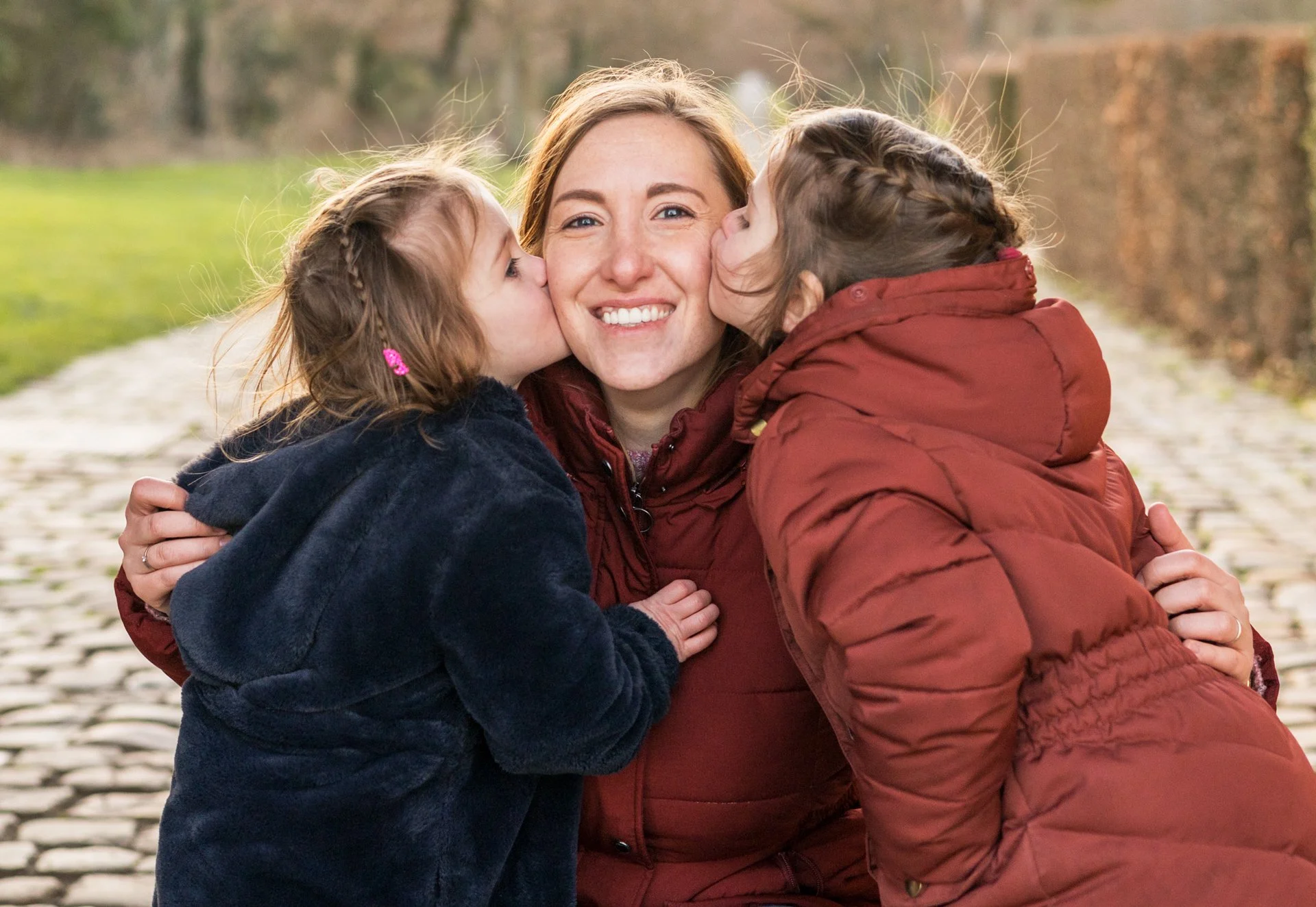 A woman with two young girls kissing her on each cheek outdoors, smiling happily on a cobblestone path lined with trees and bushes.
