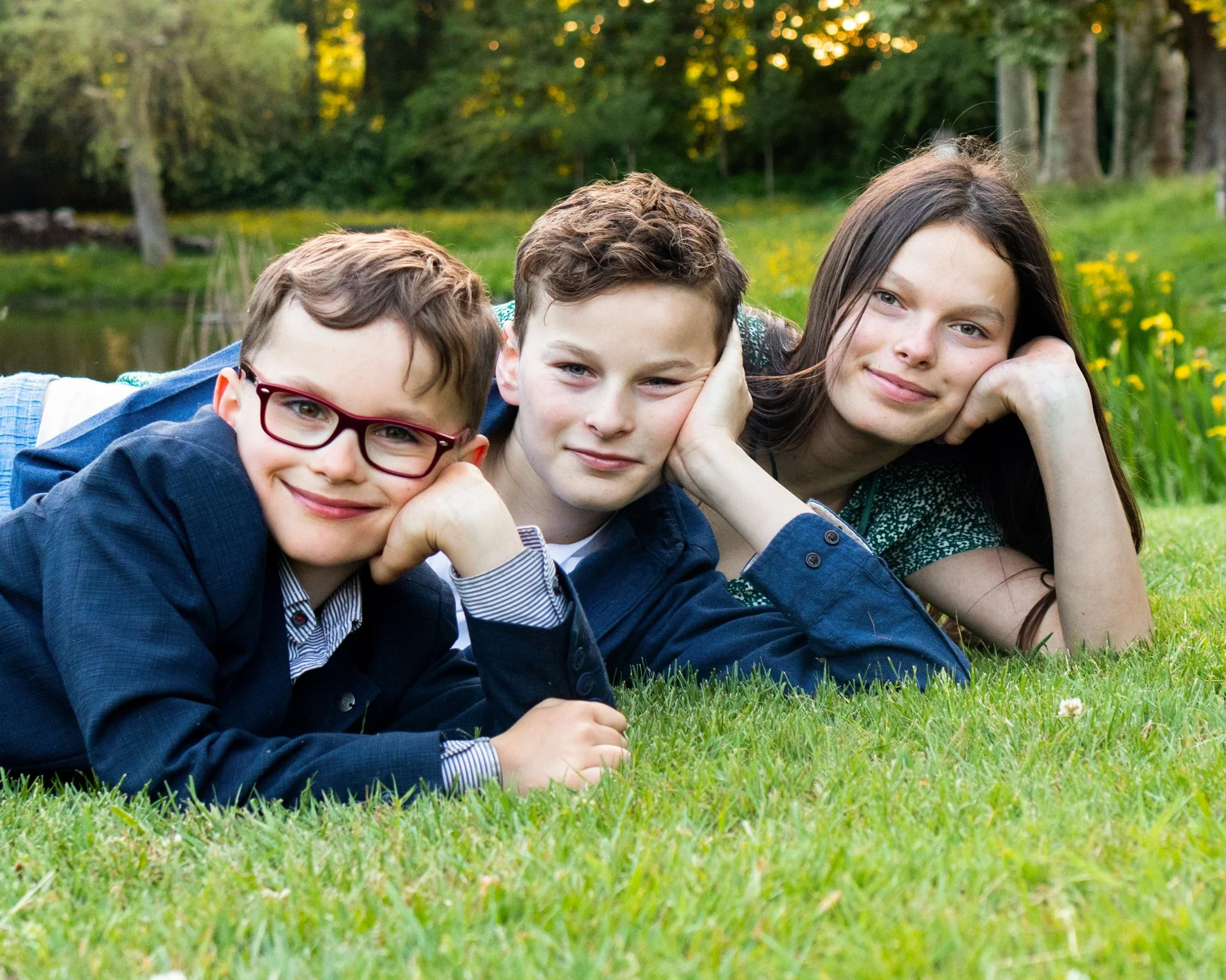 Three children lying on grass in a park, close-up, smiling, with trees and a pond in the background.
