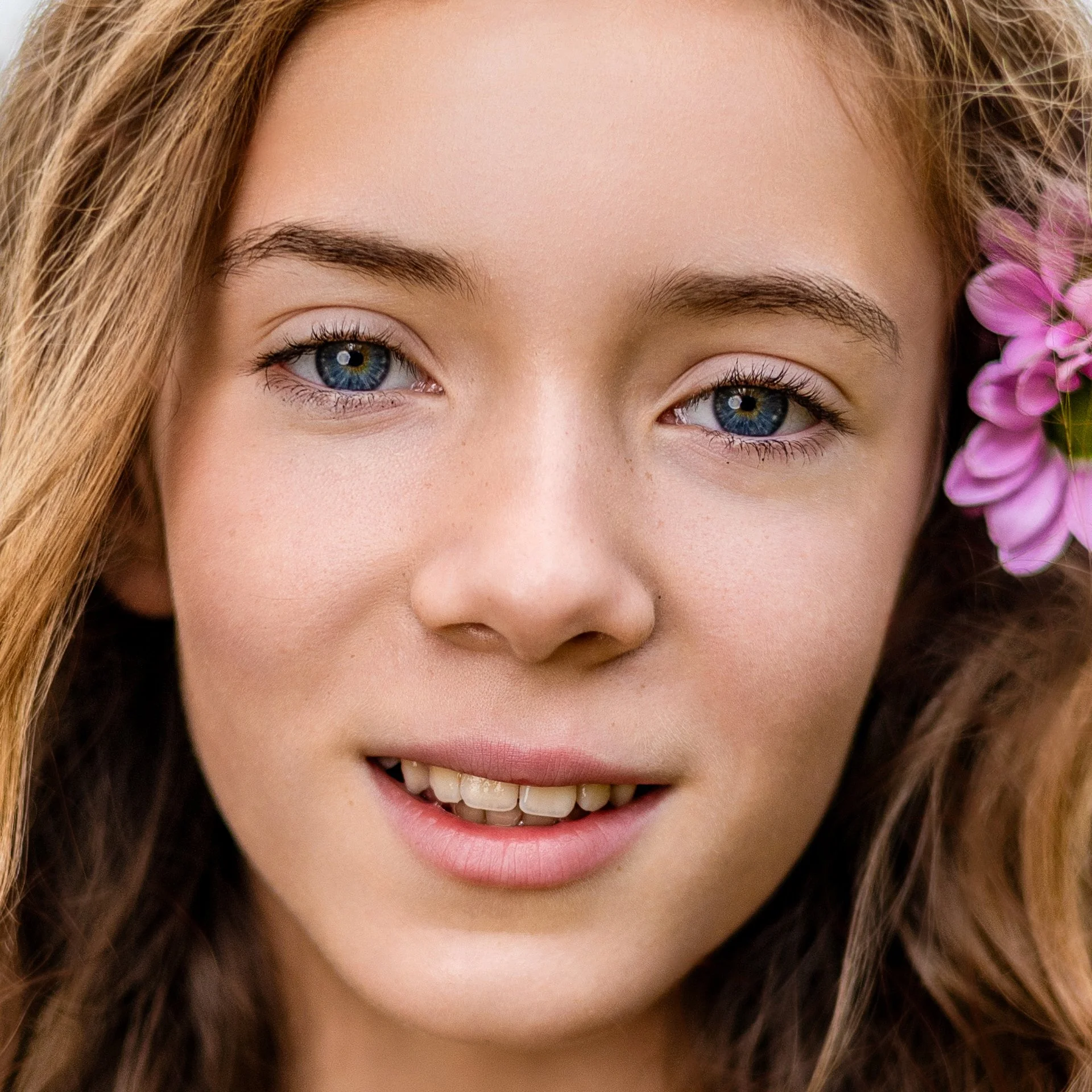 Close-up of a young woman with blue eyes, light skin, and long, wavy, light brown hair, with a purple flower near her ear.