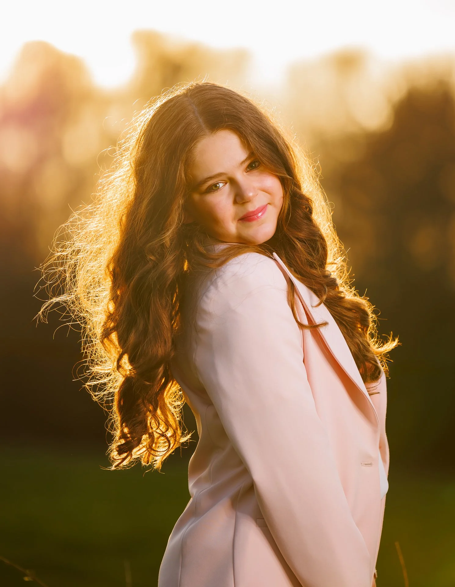 Young woman with long curly hair stands outdoors during golden hour, wearing a light pink blazer and smiling softly.