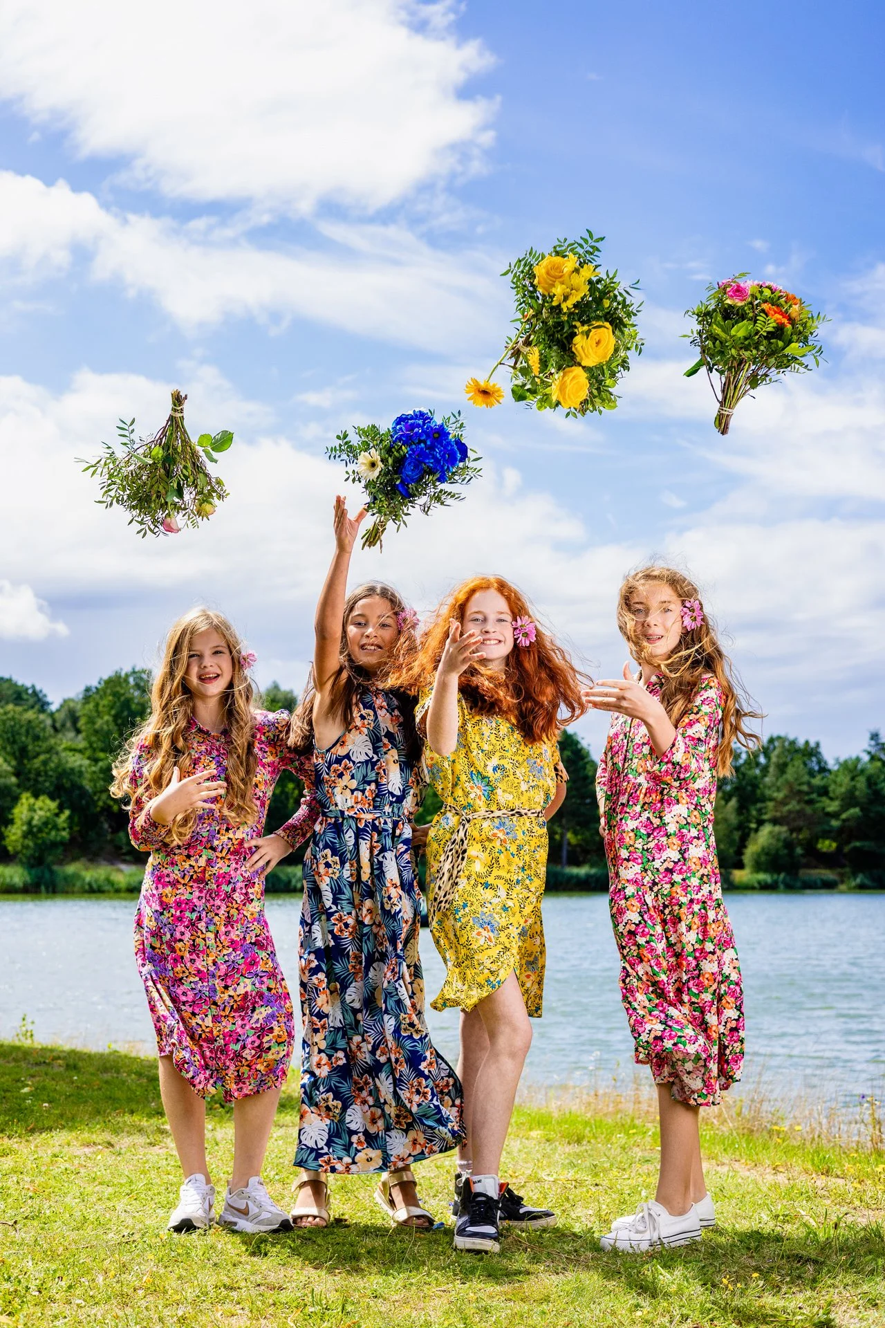 Four girls in colorful dresses by a lake, tossing bouquets of flowers into the air, under a partly cloudy sky.