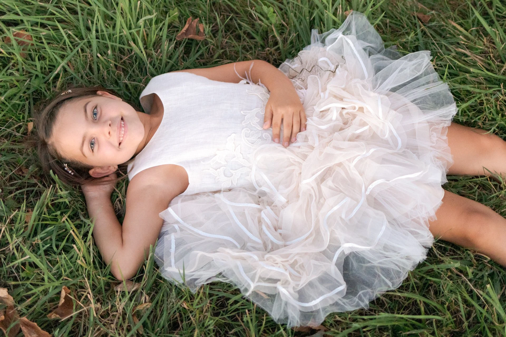 A young girl lying on grass, wearing a white sleeveless dress with a ruffled tulle skirt, smiling and looking at the camera.