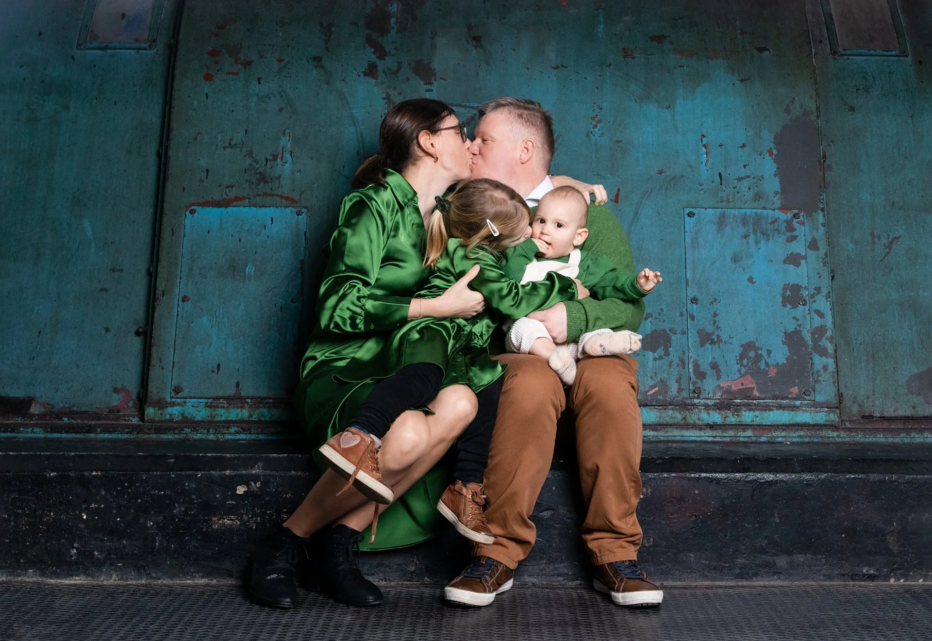 Family of four sitting against a blue-green textured wall. The parents are kissing, the mother in a green satin dress and the father in a green sweater. Their two children, a girl and a baby boy, are sitting on their laps. The girl is wearing a green