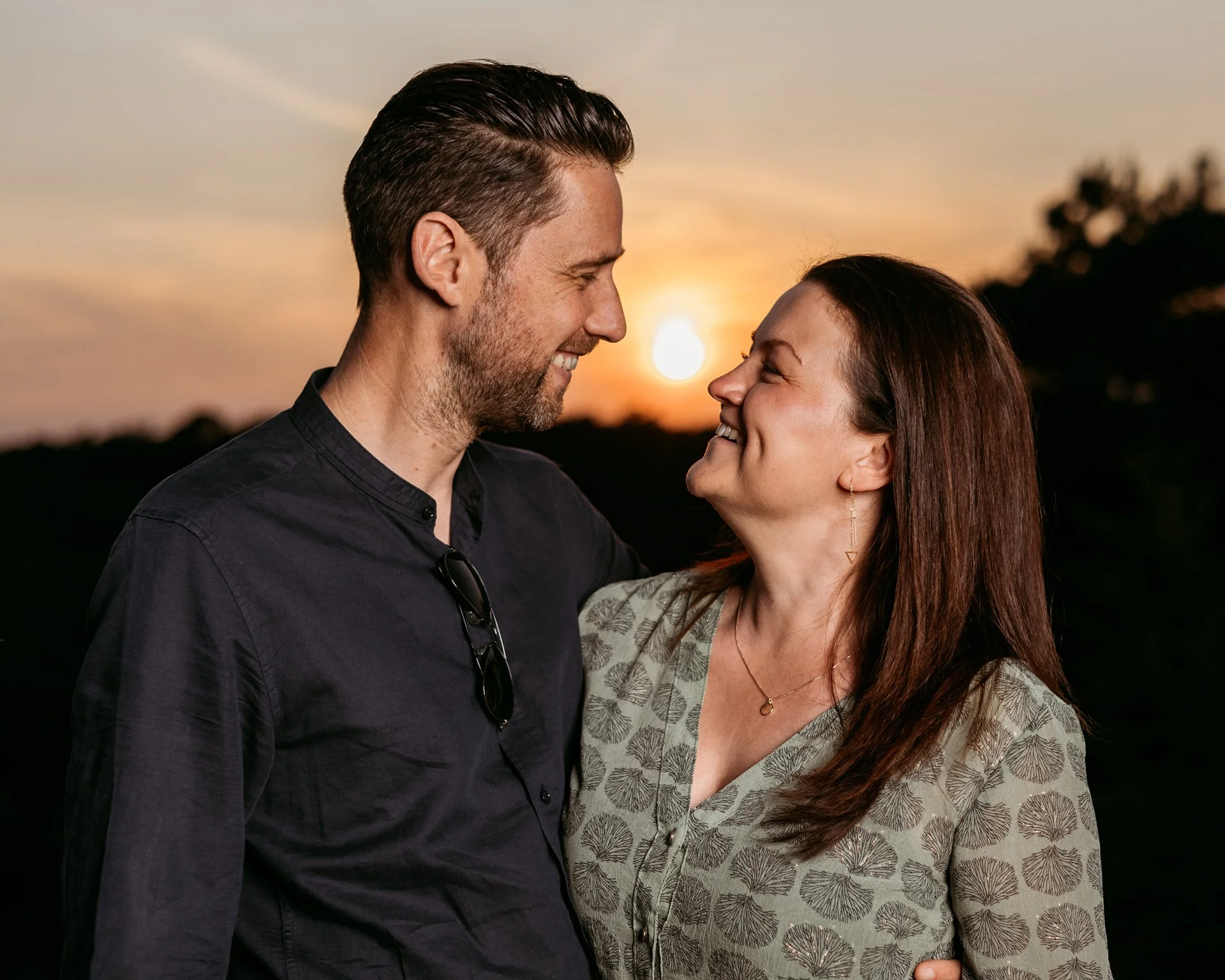 A couple smiling at each other during sunset, standing closely outdoors with a sunset in the background.