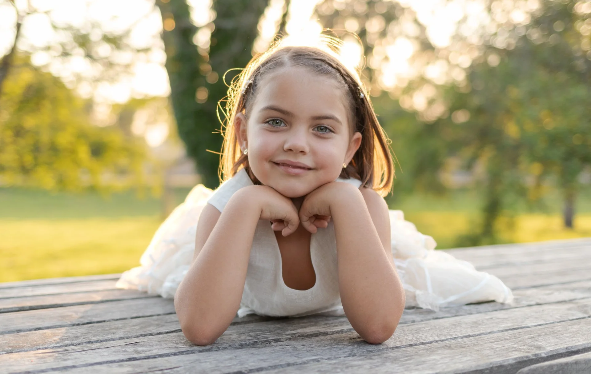 A young girl lying on her stomach on a wooden surface outdoors, resting her chin on her hands, with a background of trees and sunlight.
