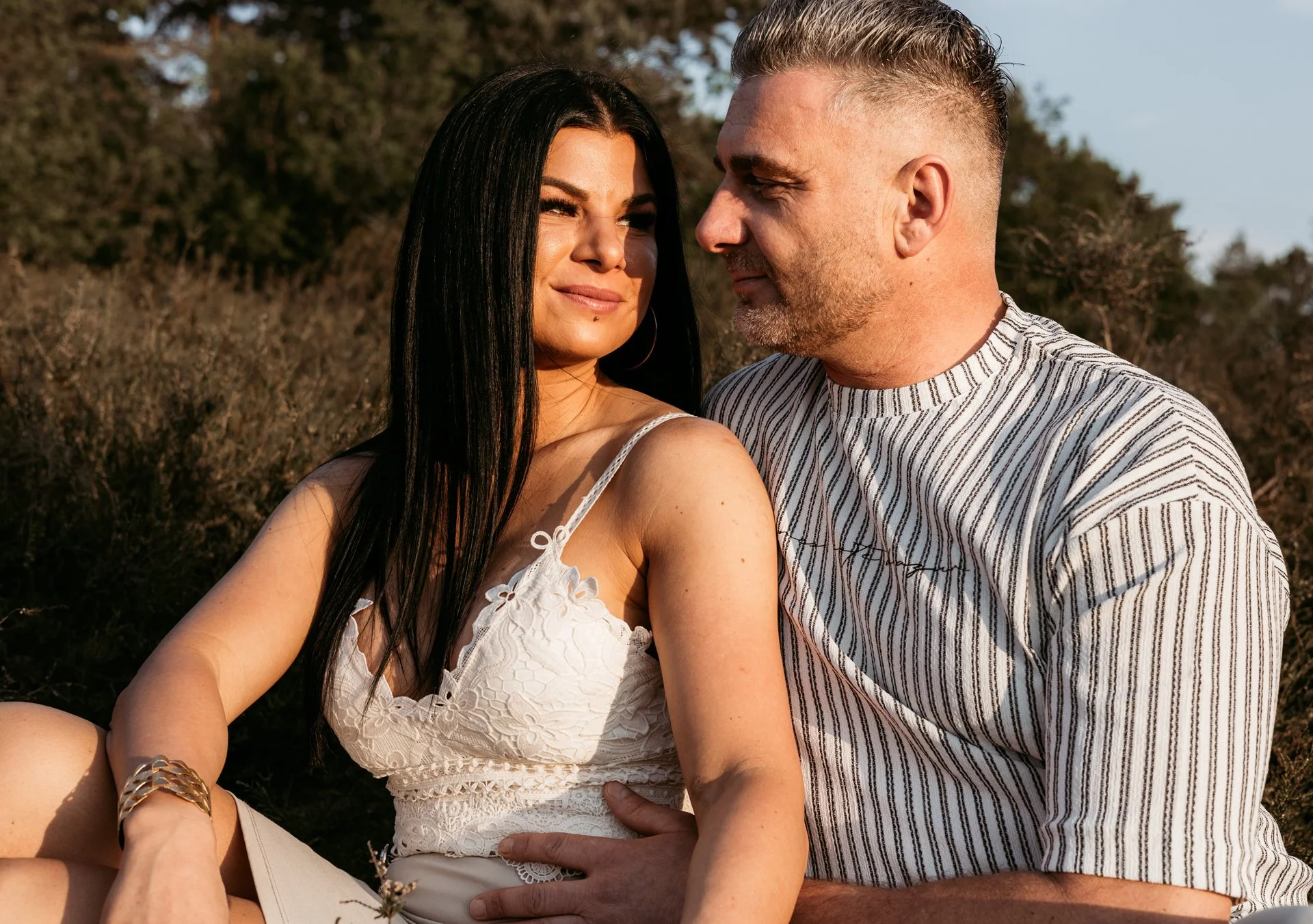 A woman and a man sitting outdoors in sunlight, looking at each other affectionately. The woman has long dark hair and is wearing a white dress with lace details, and the man has short light hair and a beard, wearing a striped shirt. They are surroun