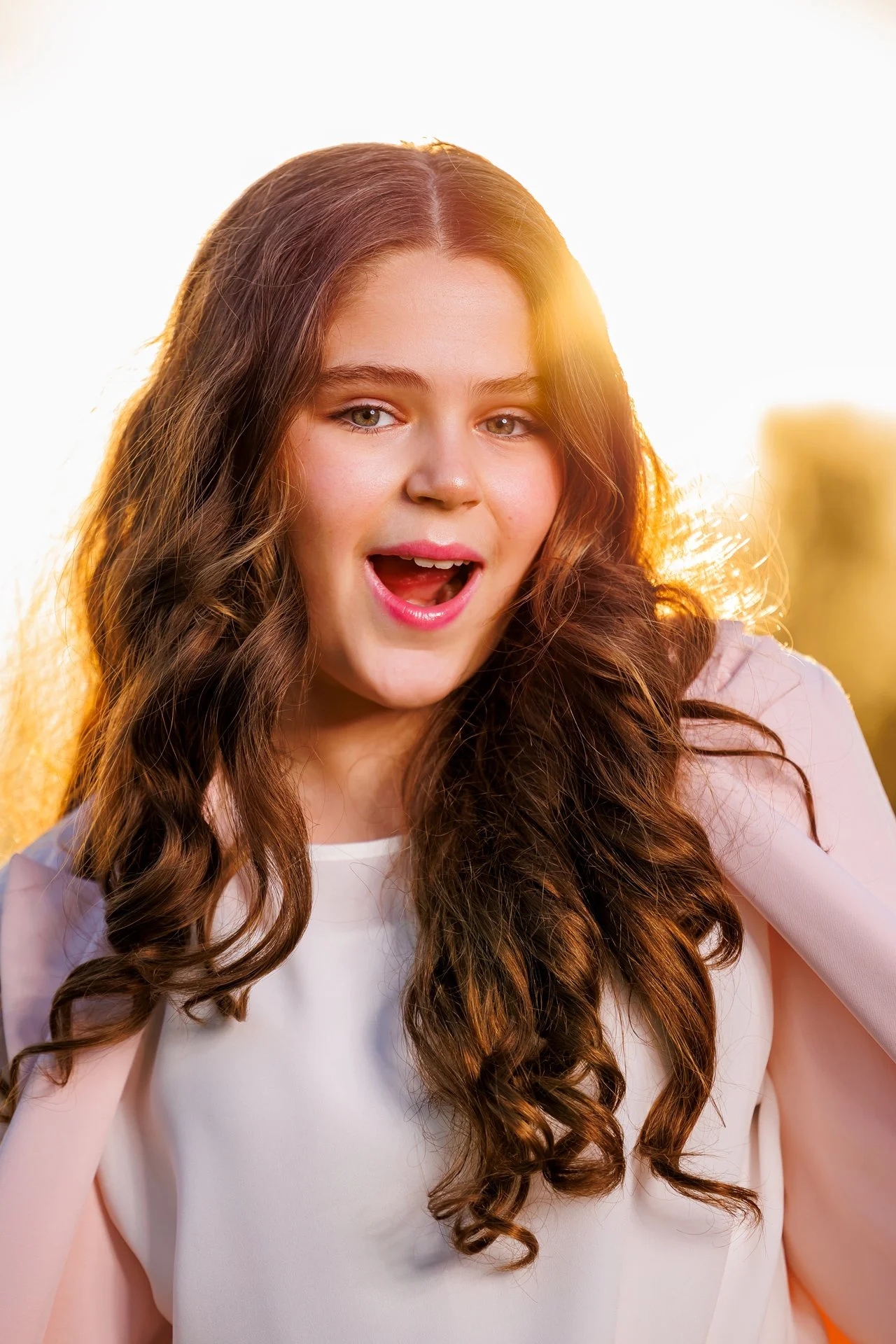 A young woman with long, curly brown hair and fair skin, smiling with her mouth open, standing outdoors during sunset.