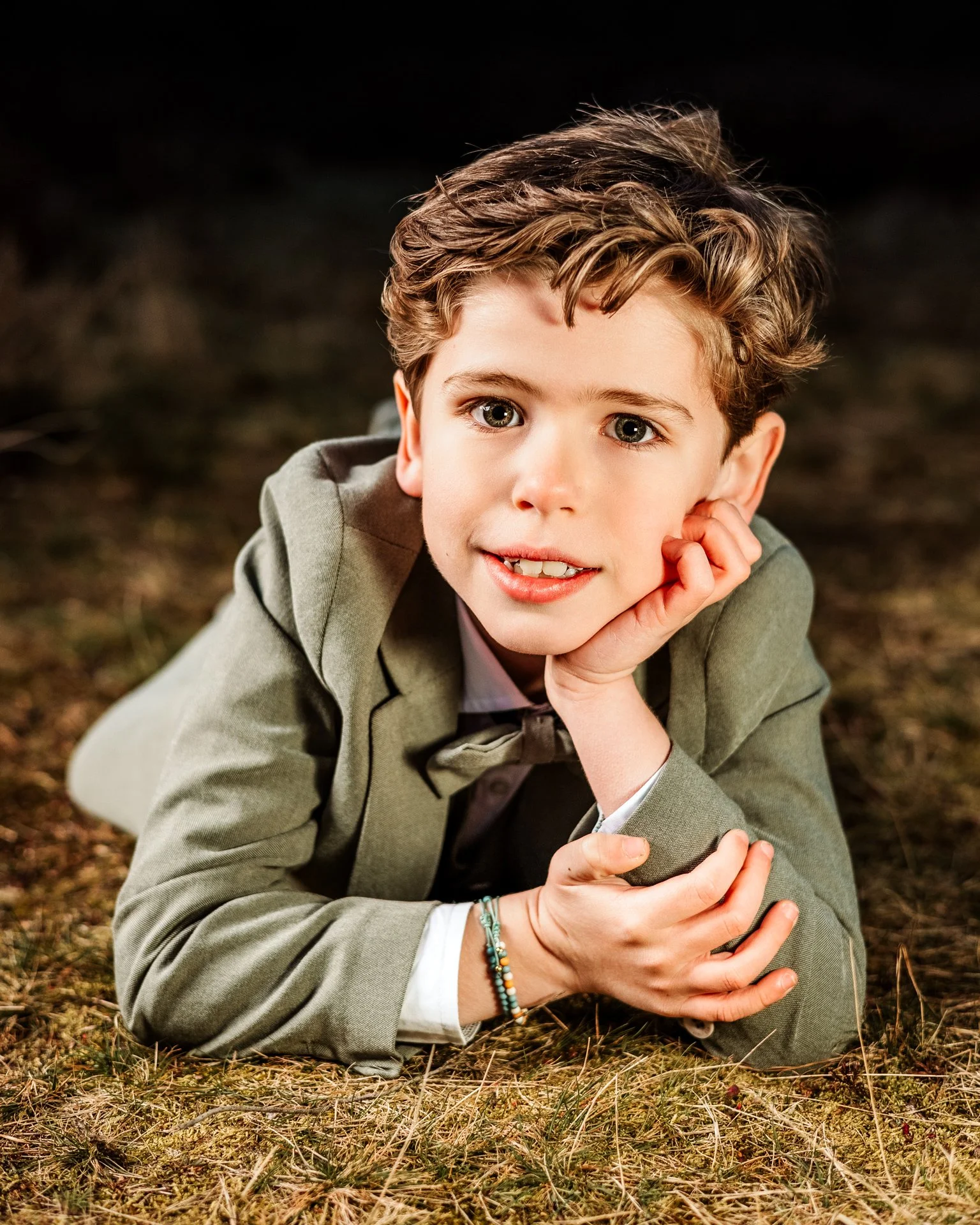 A young boy with brown curly hair and green eyes lying on the ground, resting his head on his hand, wearing a light gray suit and a white shirt, outdoors at night.
