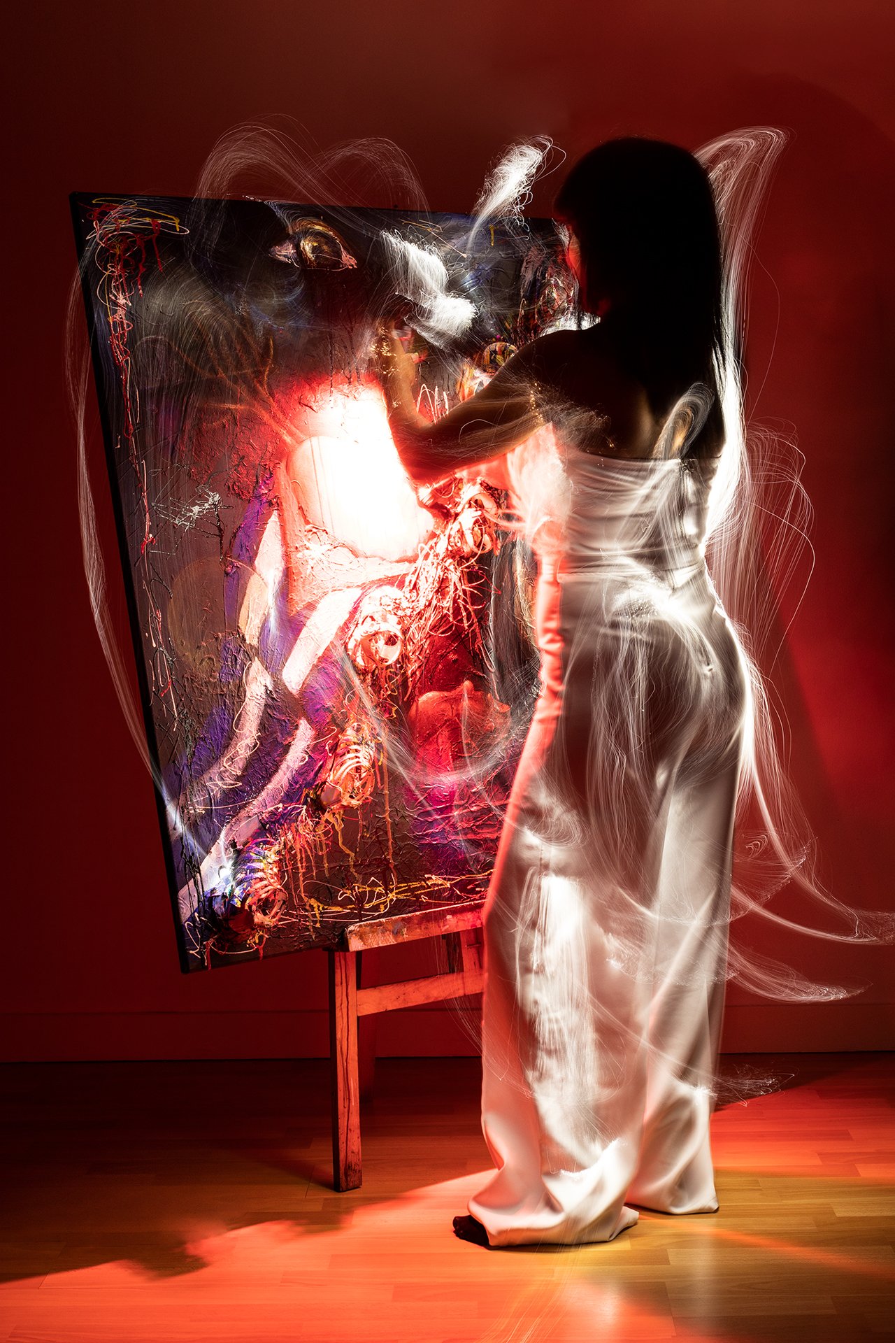 A painter in a long white dress appears to be painting on an abstract canvas with vibrant colors, with light painting trails surrounding her in a dark room.