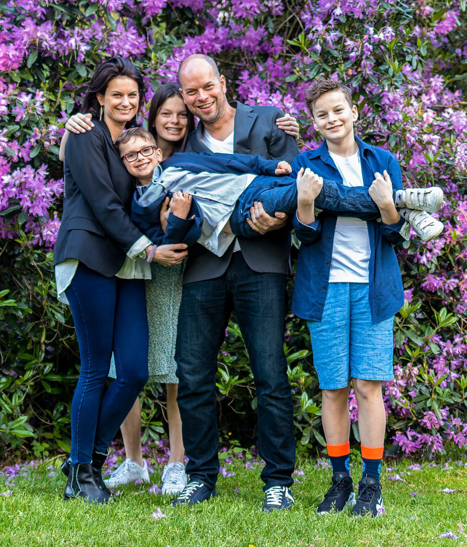 Family of six standing outdoors in front of pink flowering bushes, with a man holding a child and a boy giving a thumbs up.