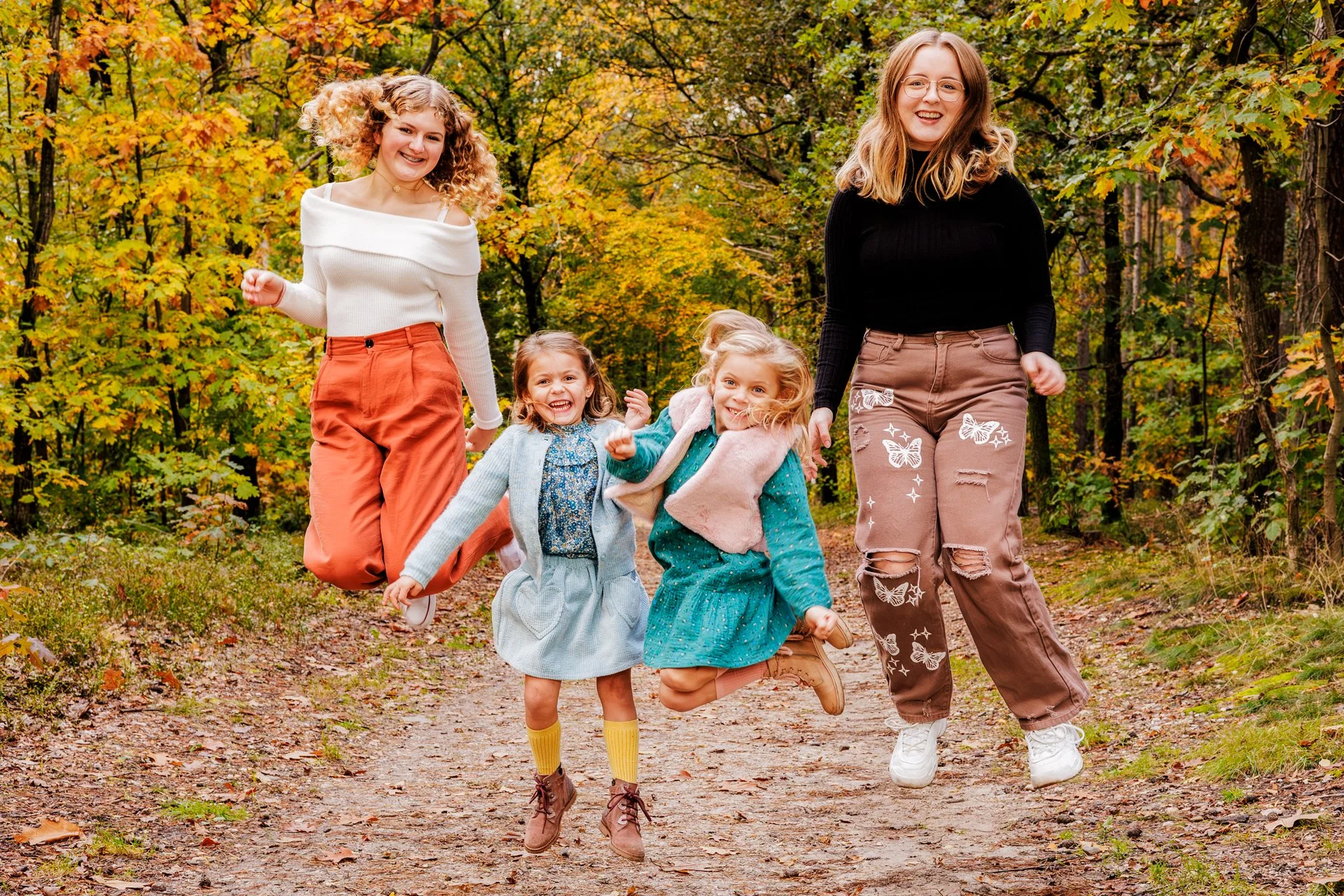 A group of five women, including three young girls and two adult women, joyfully jumping on a forest trail during autumn, surrounded by trees with colorful fall foliage.