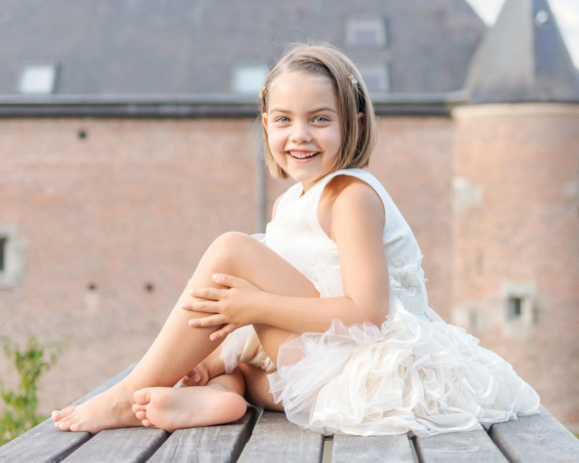 A young girl sitting on a wooden surface outdoors, wearing a white dress, smiling, with a brick building in the background.