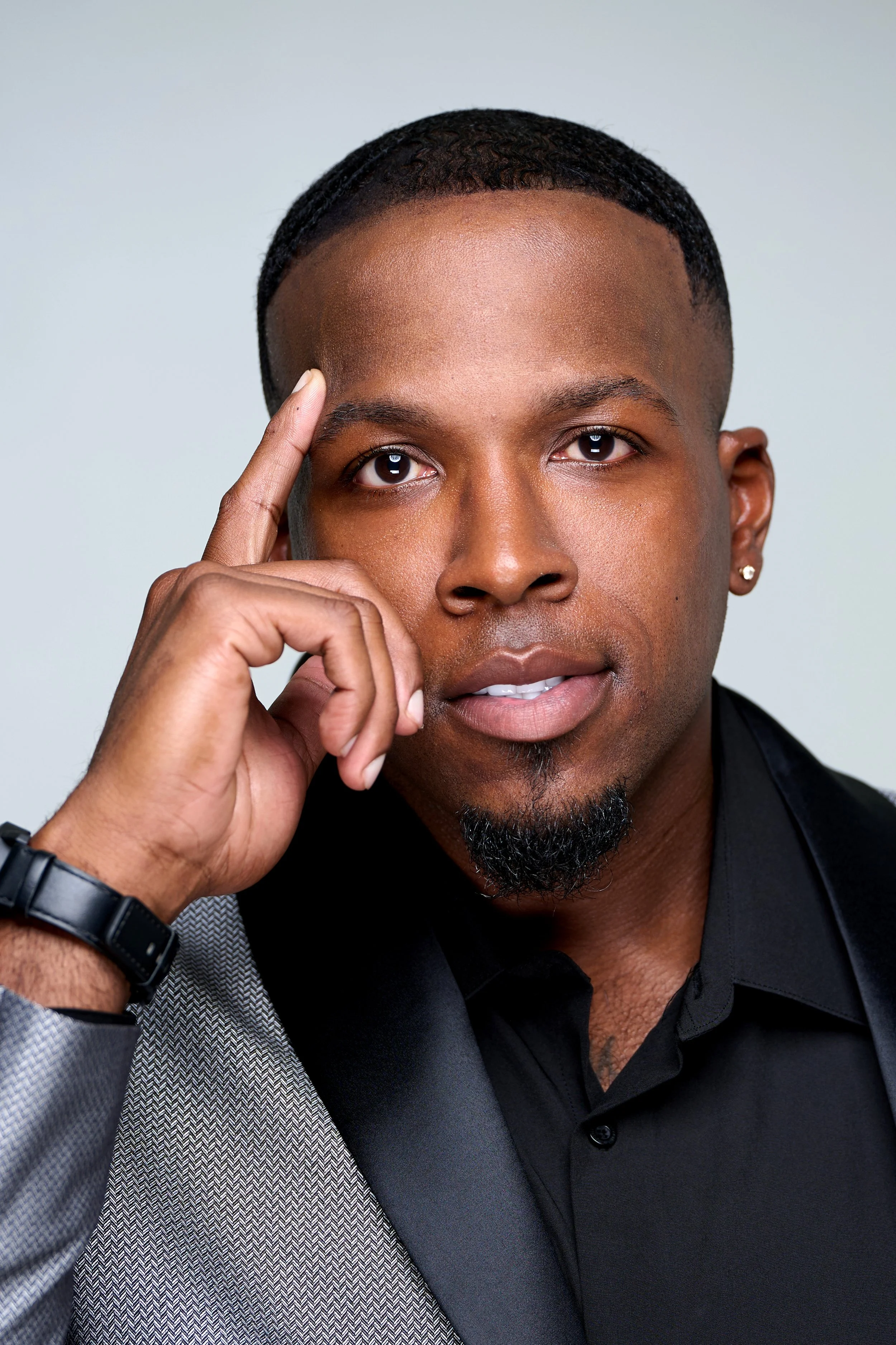 Close-up of a young African American man with short hair, dressed in a black shirt, wearing a gray blazer, and making a gesture with his right hand near his temple. He has a confident expression and is looking directly at the camera against a plain, light background.