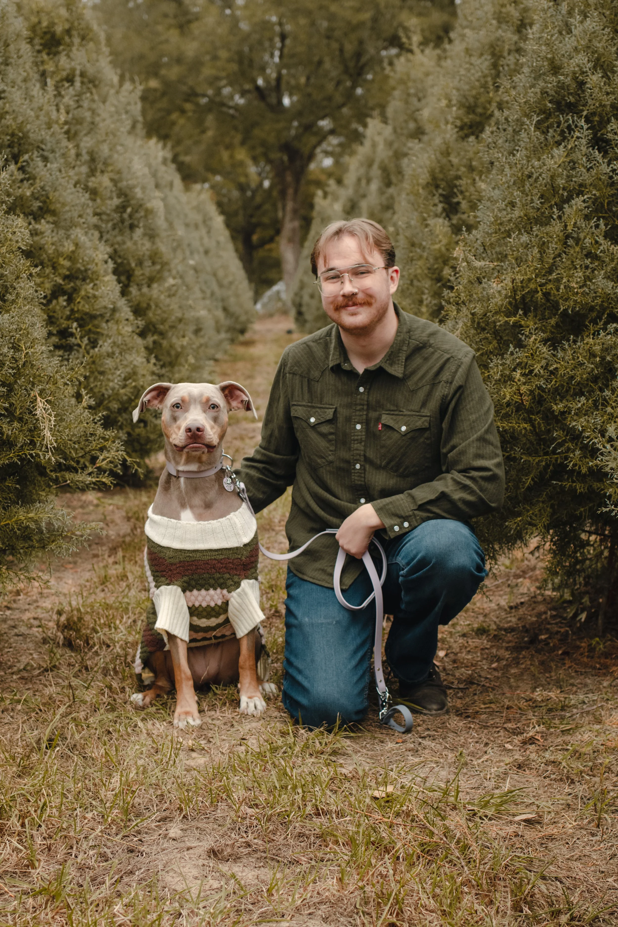 A man with glasses and a beard kneeling with a dog in an outdoor setting surrounded by bushes and trees, both looking at the camera.