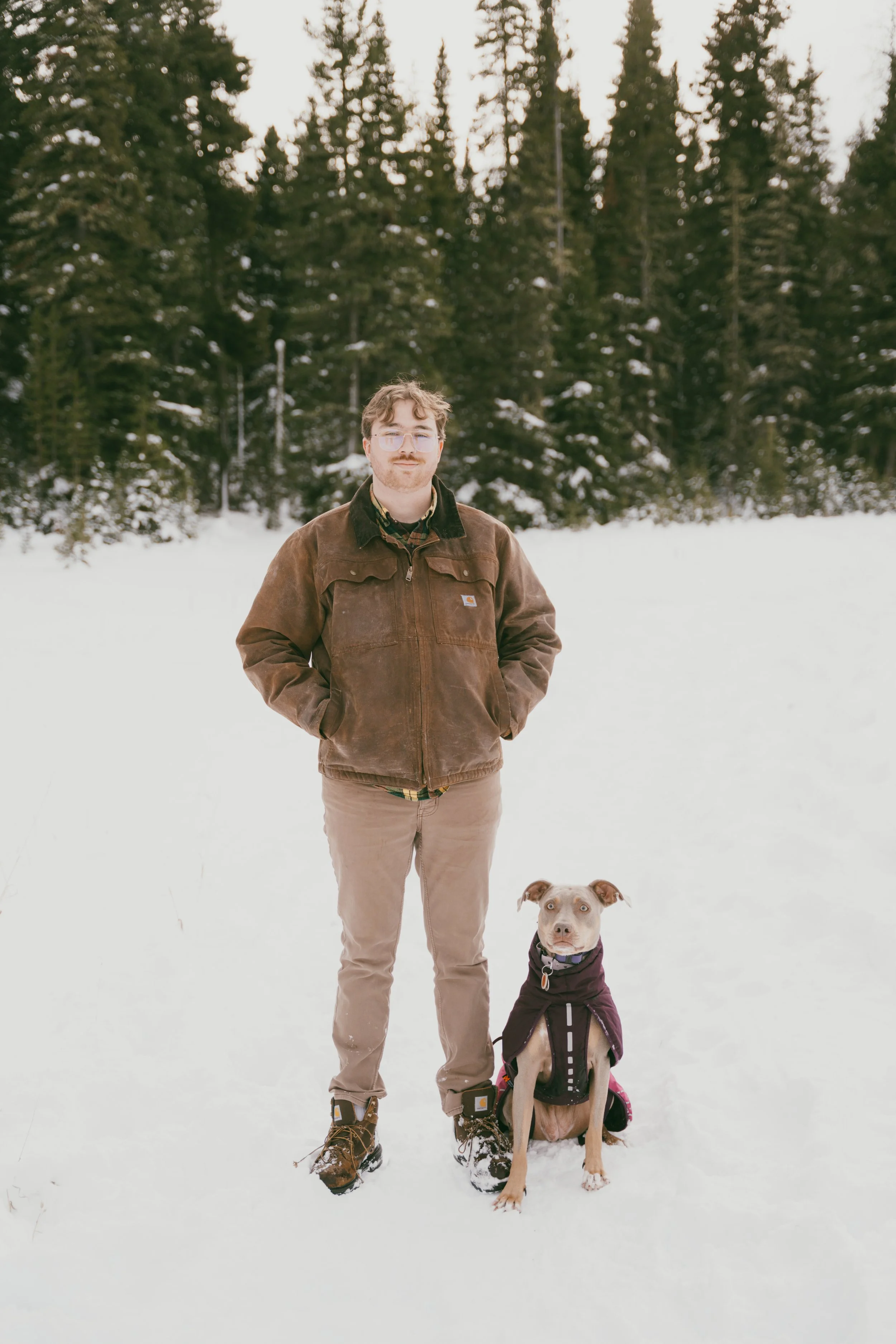 A man standing in the snow with a dog sitting beside him, surrounded by a forest of evergreen trees.
