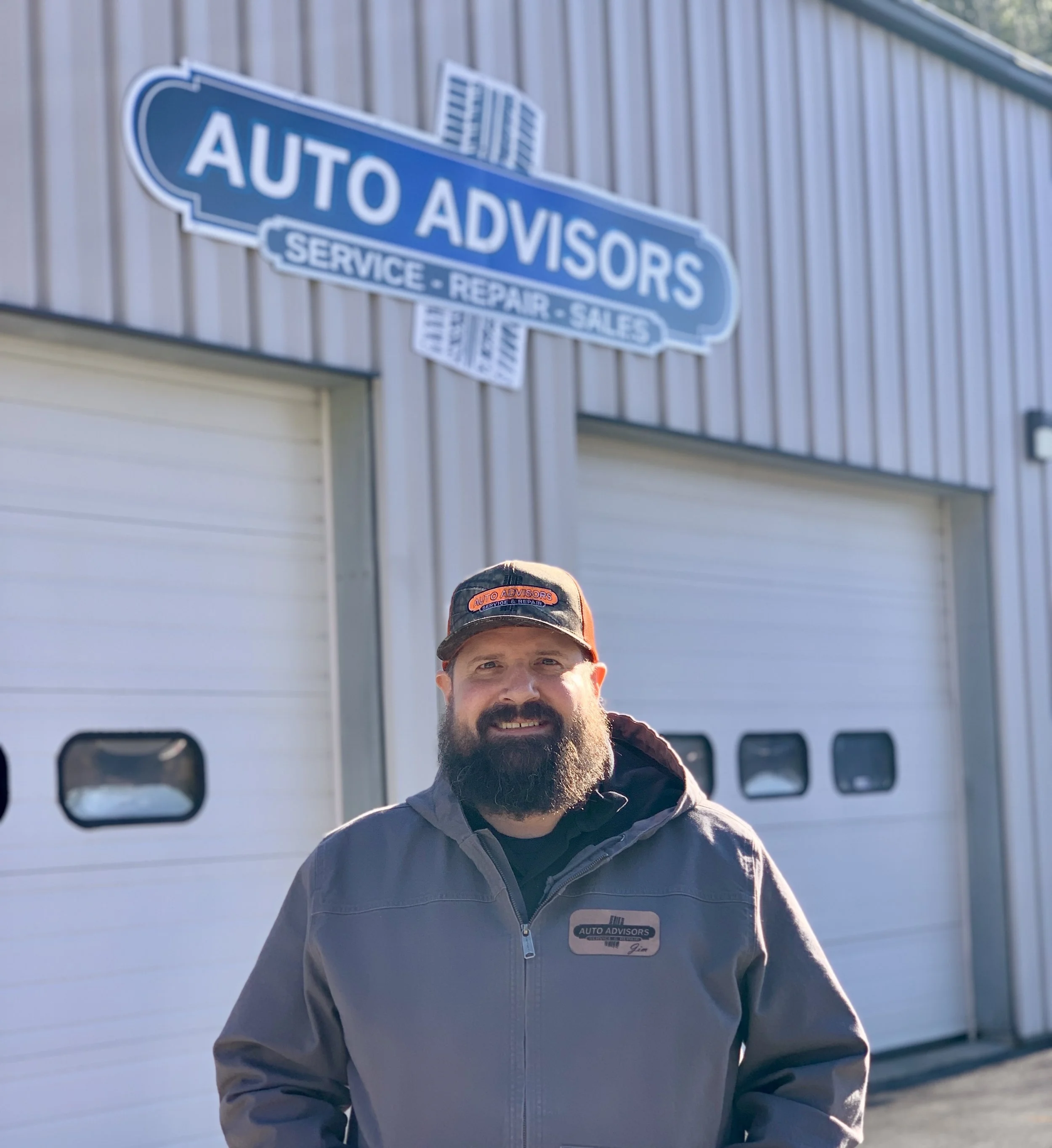 A smiling man with a beard wearing a black and blue work uniform with the name tag 'Jim' and an 'Auto Advisors' patch, inside a garage or auto repair shop with tires and automotive equipment in the background.