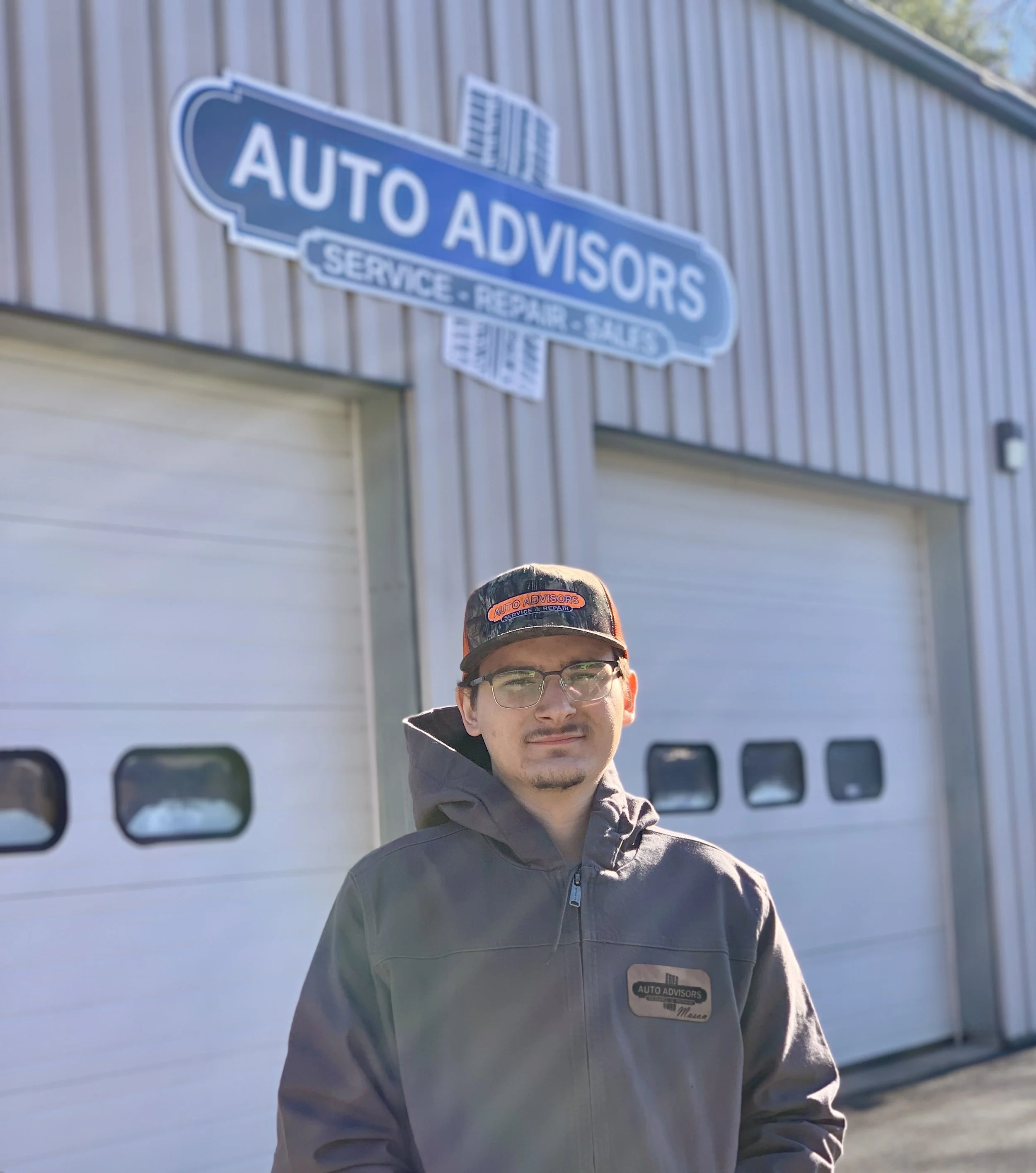 Young man with glasses and a cap smiling, wearing a navy sweatshirt with 'Auto Advisors' logo, standing indoors against a wall painted beige, red, and blue.