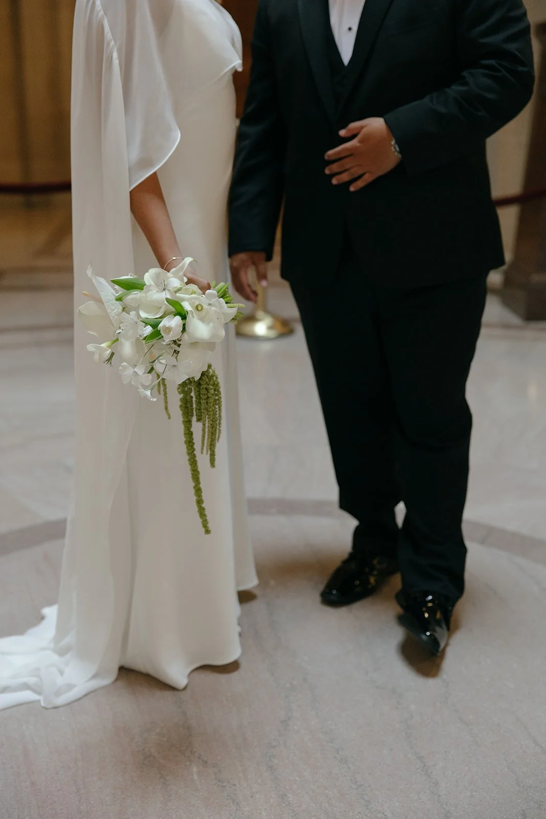 A bride and groom holding hands during their wedding ceremony. The bride is holding a bouquet of white flowers and wearing a white dress. The groom is dressed in a black tuxedo.