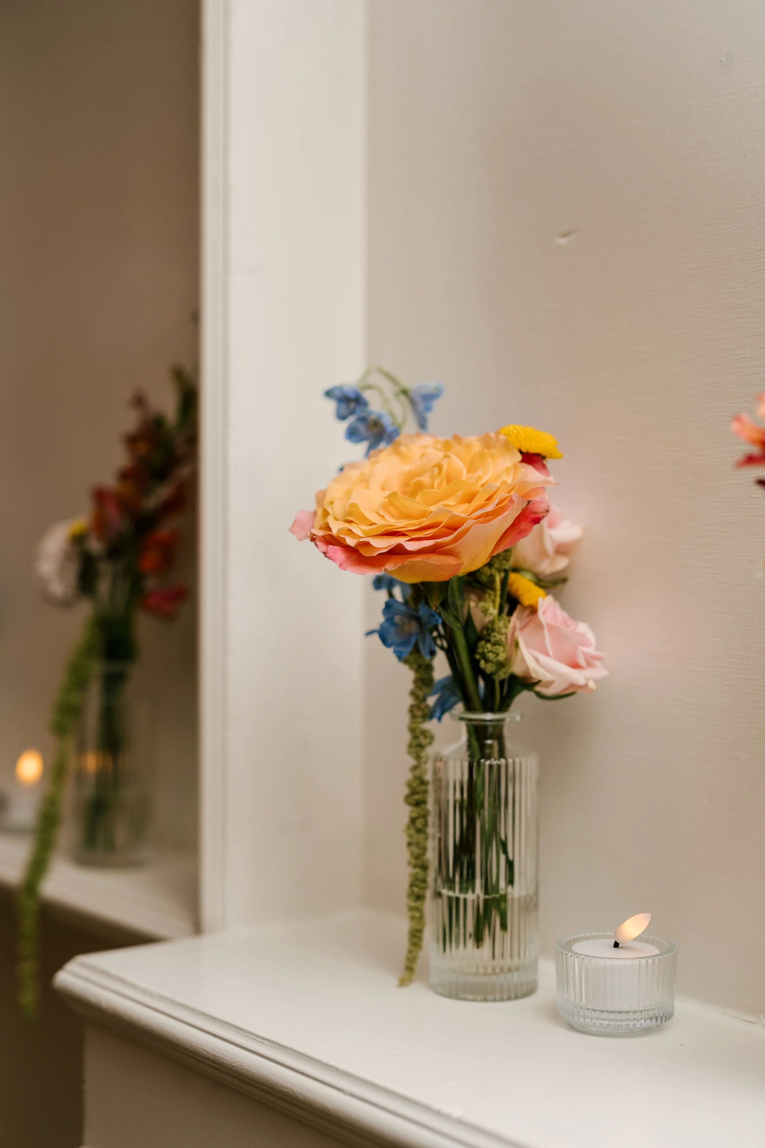 A vase with a bouquet of pink, yellow, and blue flowers on a white surface, with a mirror and a lit candle in a clear glass holder beside it.