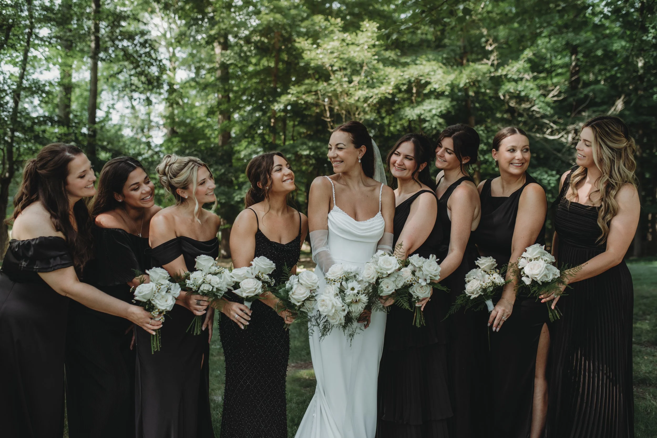 A bride in a white dress and veil standing amidst eight bridesmaids in black dresses, all holding white flower bouquets, in a lush green outdoor setting during daytime.