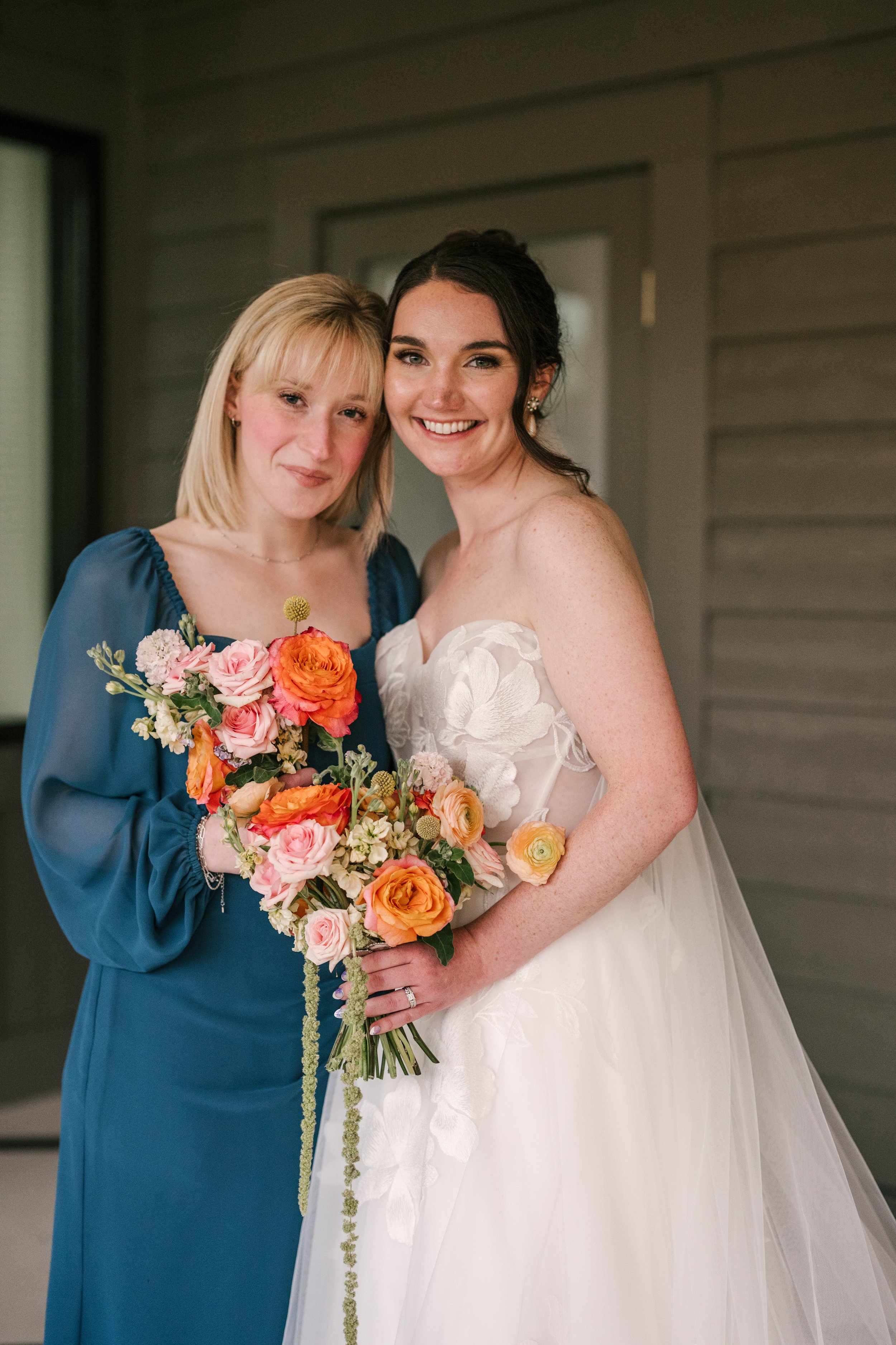 Two women, one in a white wedding dress and the other in a blue dress, standing close together and smiling, holding a large bouquet of pink, peach, and white flowers.