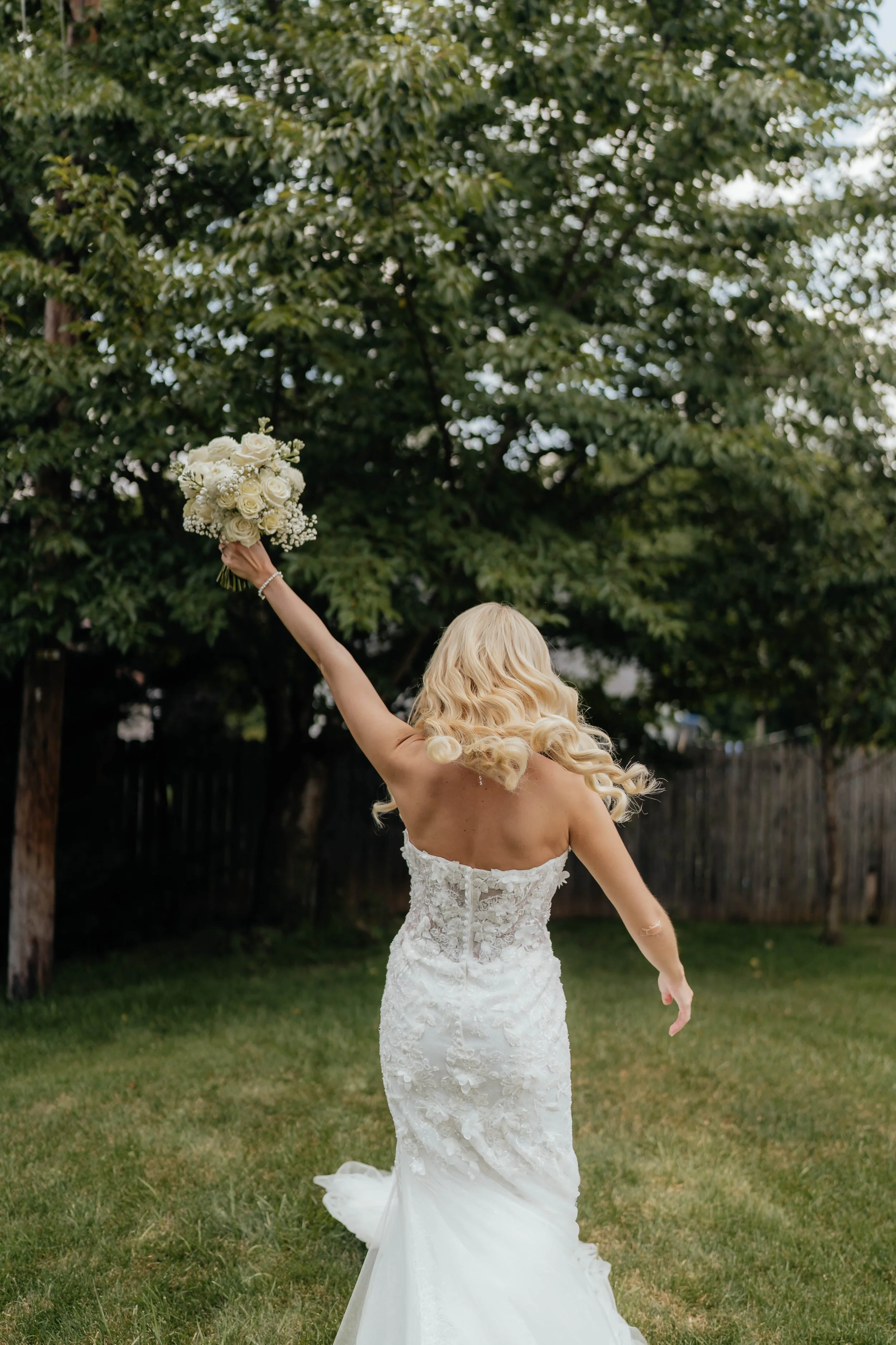 A bride in a lace wedding dress holding a bouquet of white roses and baby's breath, raising her arm outdoors with trees and a wooden fence in the background.