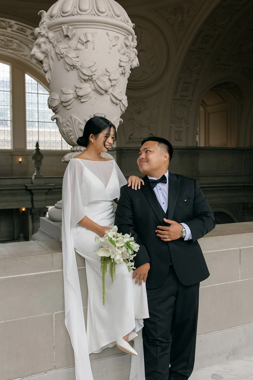 A bride in a white wedding dress and a groom in a black tuxedo with a bow tie pose together inside a grand, historic building with large windows and ornate architectural details. The bride is holding a bouquet of white calla lilies and smiling at the groom, who is looking at the bride with a smile.