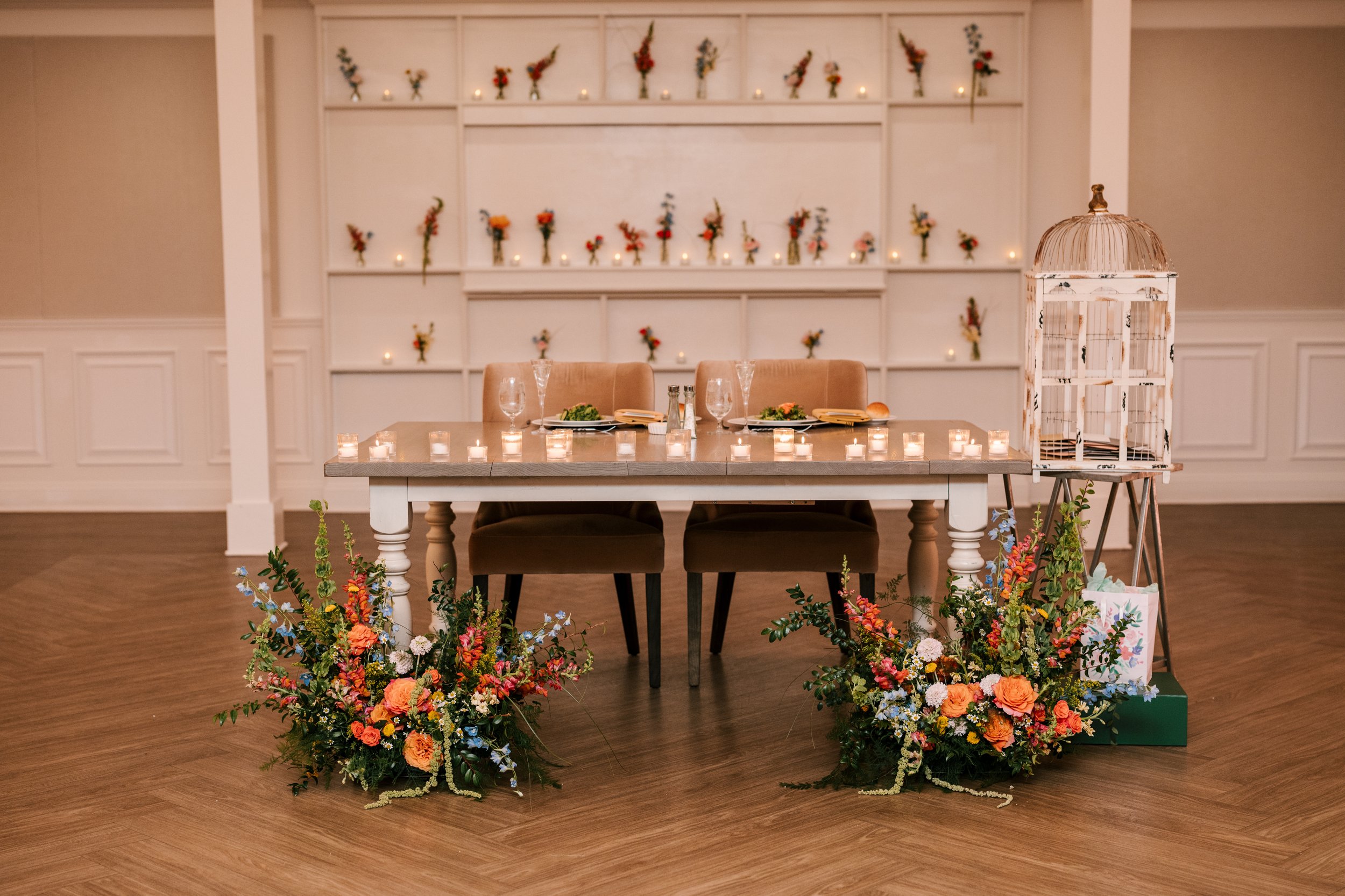 A decorated table with plates and glasses, surrounded by colorful floral arrangements and candles, set in a room with a wall of small flower bouquets and candles in the background for an event or celebration.
