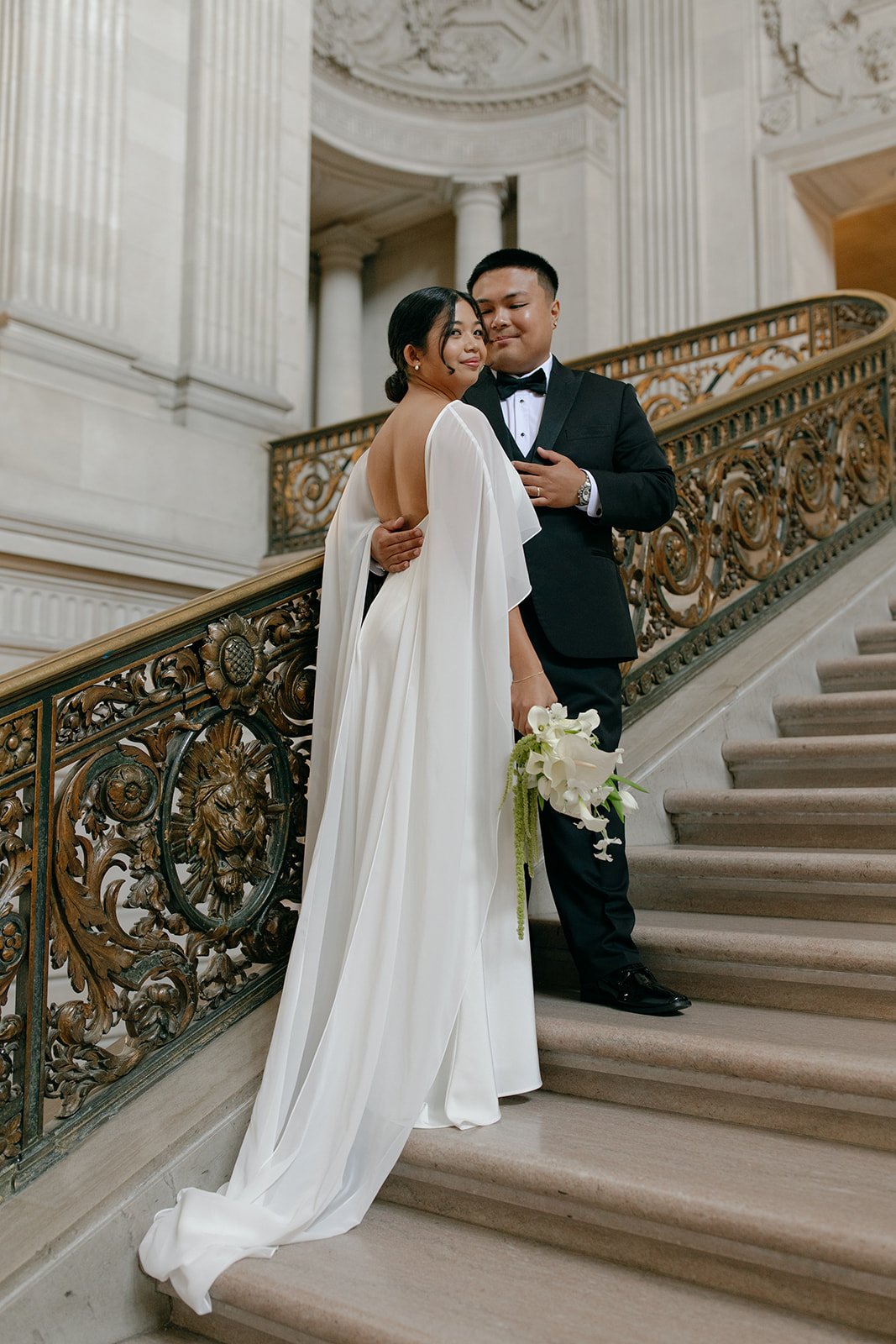 A bride and groom standing on an ornate staircase inside a grand building during a wedding ceremony. The bride is wearing a white gown and holding a bouquet of white flowers, while the groom is in a black tuxedo. They are close together, sharing an intimate moment.