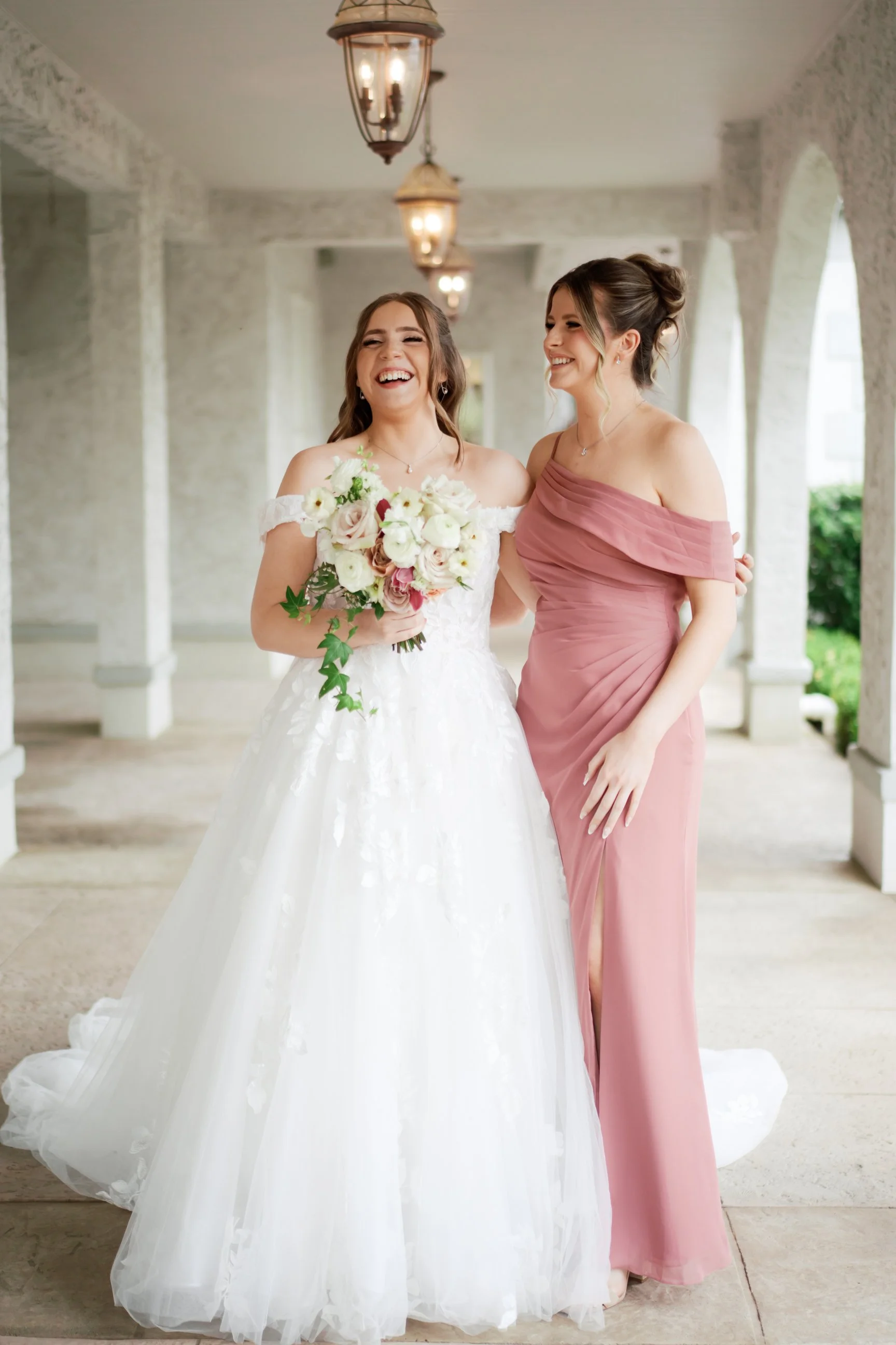 Two women, one in a wedding gown holding a bouquet, and the other in a pink dress, standing together and smiling in a bright, arched hallway with hanging lantern lights.