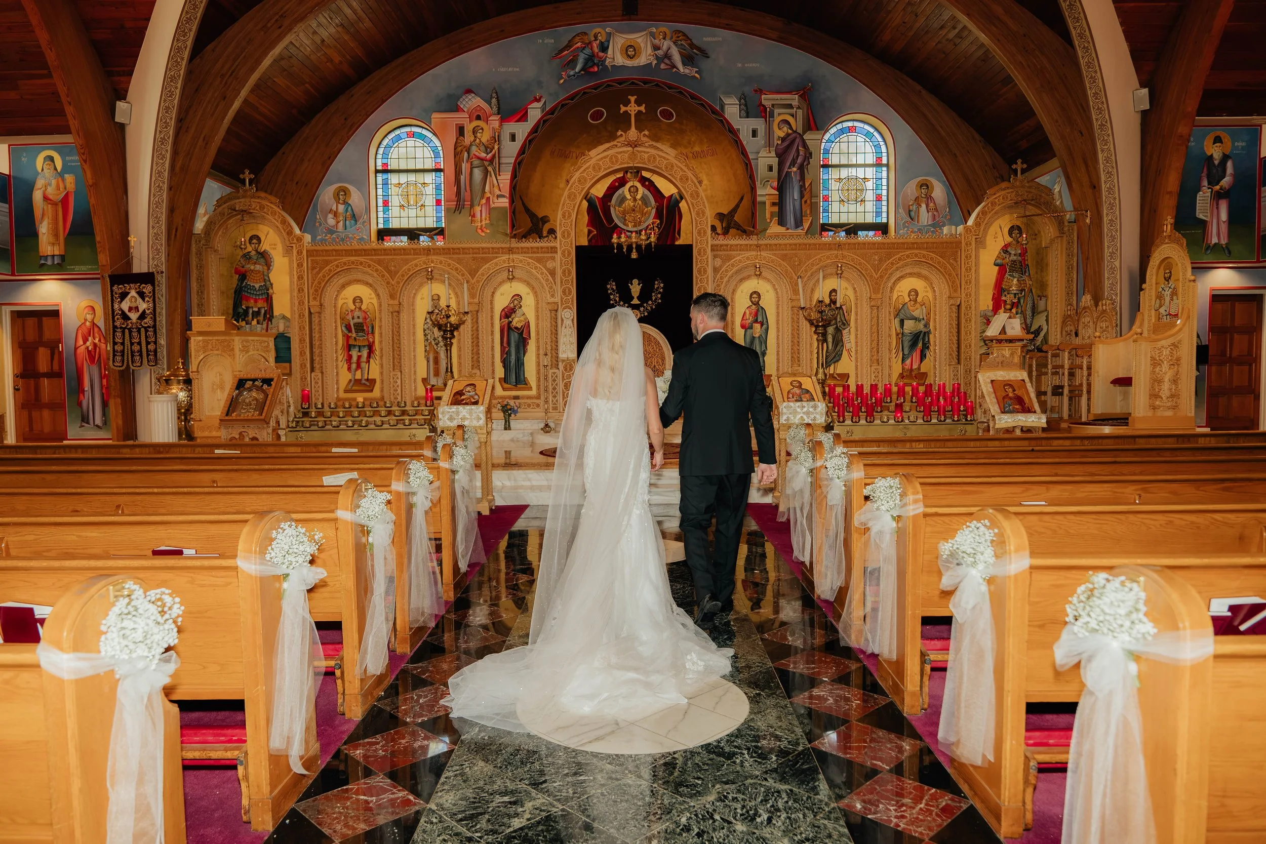 A bride and groom holding hands standing at the altar of a church during their wedding ceremony. The church interior features wooden walls, ornate religious iconography, stained glass windows, and decorated pews with white flowers.