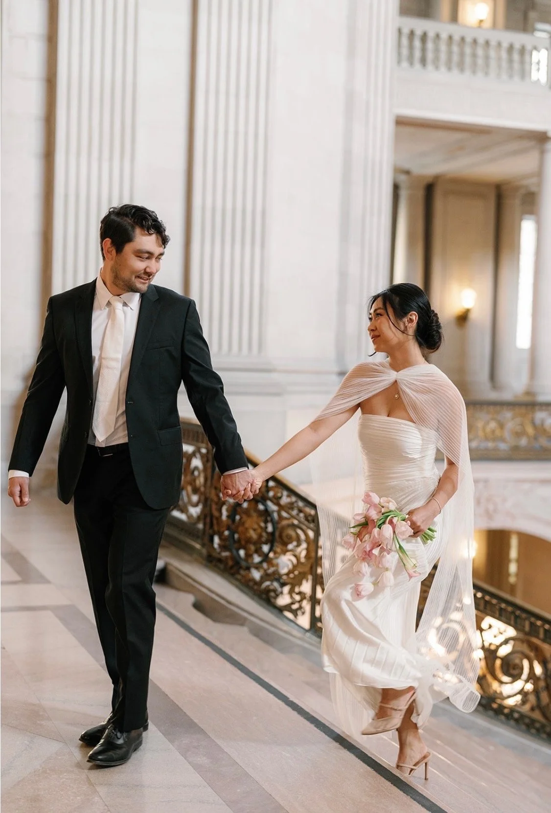 A bride and groom holding hands and walking in an elegant, grand hallway with ornate railings and tall columns.
