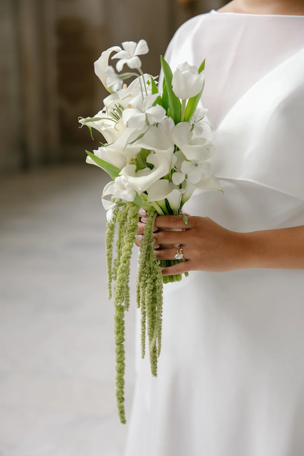 Person in white dress holding a bouquet of white flowers, including lilies, roses, and greenery with long hanging green accents, against a blurred background.