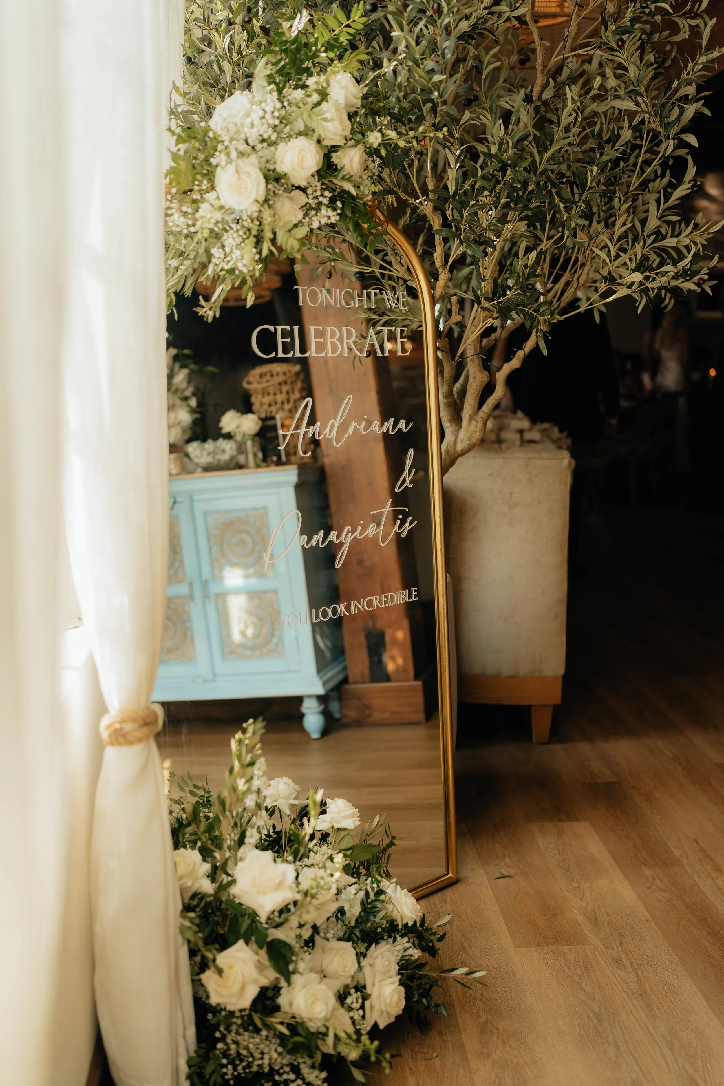 A mirror with a gold frame leaning against a wall, decorated with white flowers and greenery, with a sign that says 'Tonight we celebrate' and the names 'Andriana & Panagiotis' reflecting on the mirror. Nearby is a cream-colored curtain tied with a knot and a chest of drawers painted light blue with ornate gold accents.