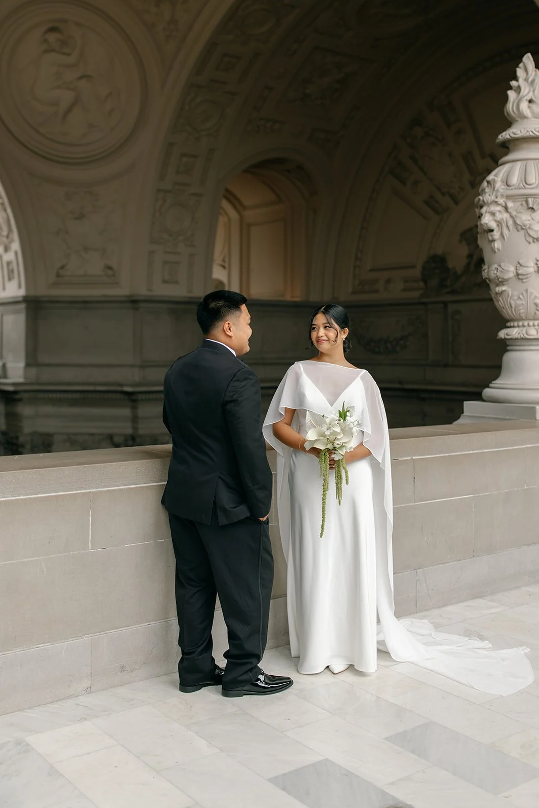 A bride and groom standing together in an elegant, ornate indoor setting with intricate ceiling decorations. The bride wears a white gown and holds a bouquet, while the groom wears a black suit. They look at each other affectionately.