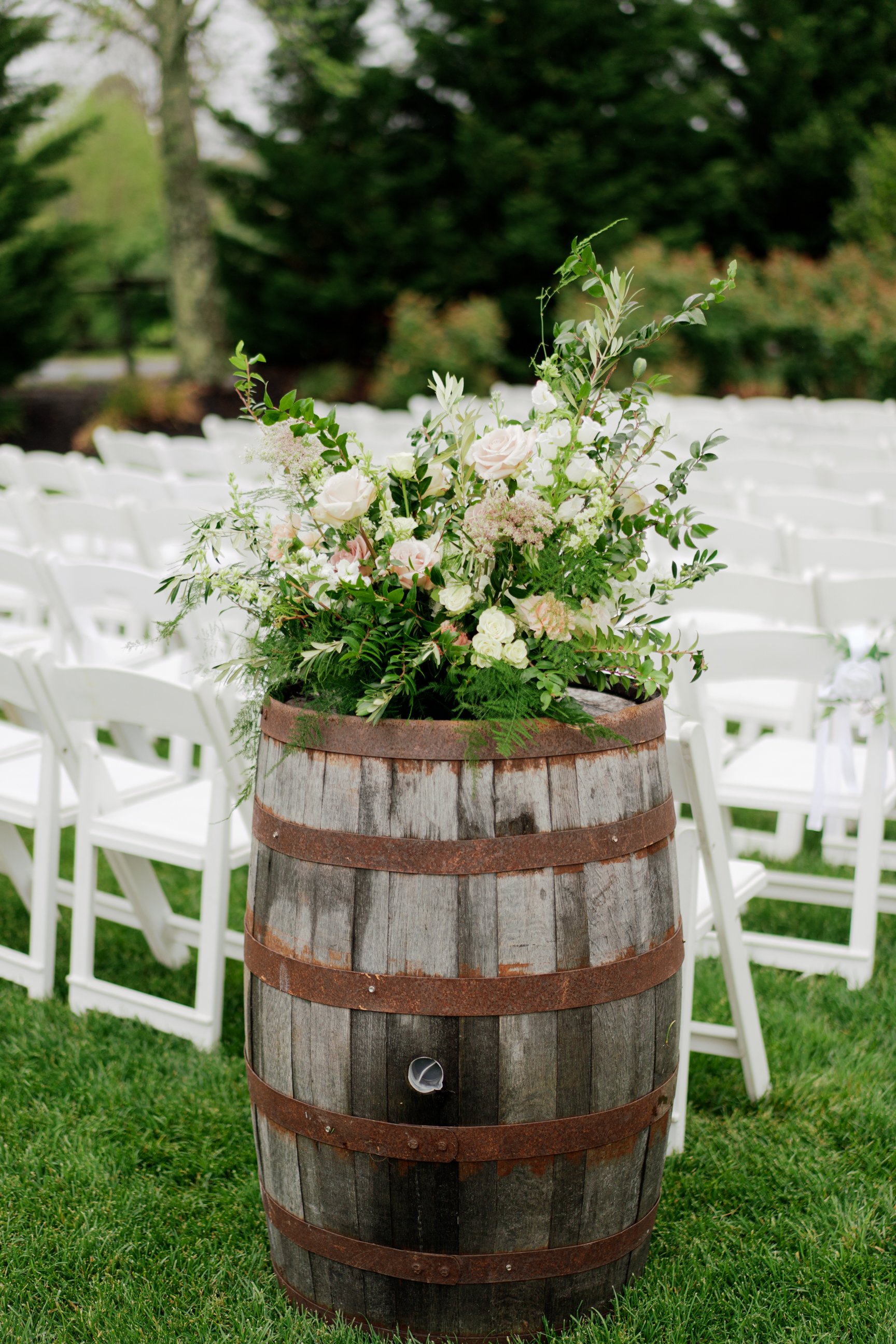 A floral arrangement with white and pink flowers and green foliage placed on top of a wooden barrel at an outdoor event with white chairs and green grass.
